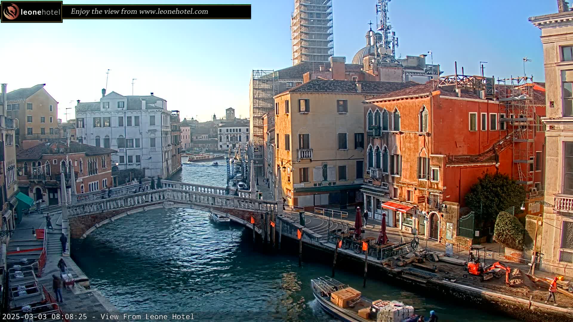 A sunny view of a canal in Venice, Italy, showing a stone bridge and colorful buildings, with some construction equipment visible along the canal's edge.