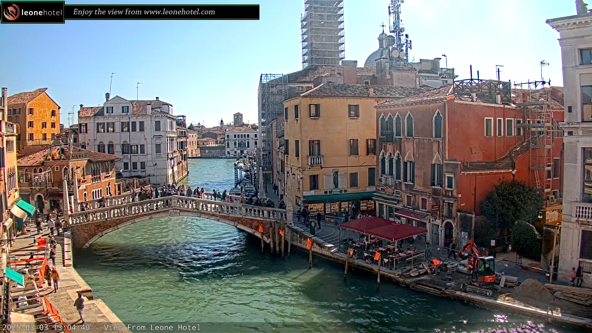 A sunny day in Venice shows a canal with a stone bridge crowded with people,  old buildings lining the waterway, and construction equipment on the shore.