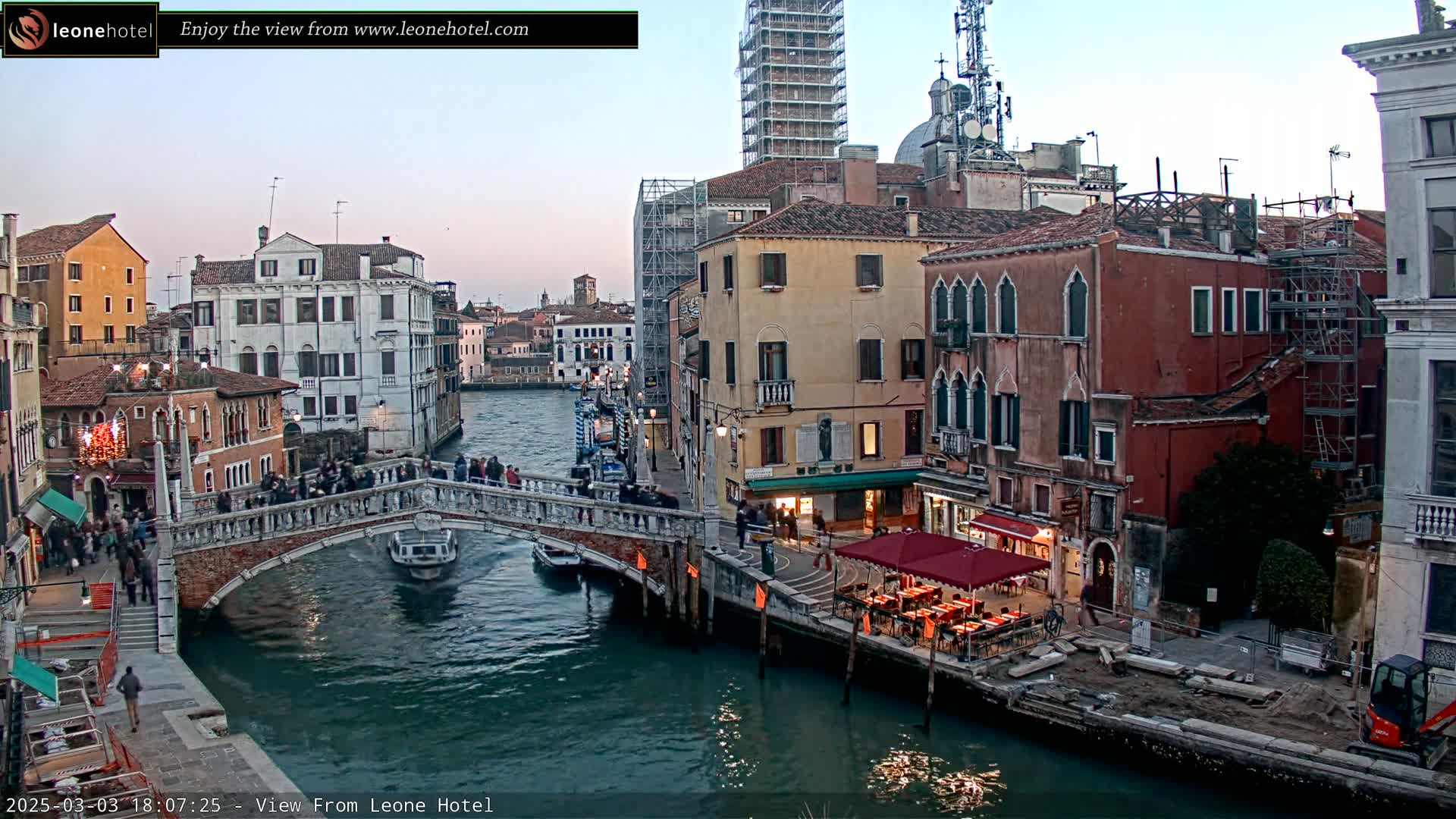 A stone bridge crosses a canal in Venice, Italy, at dusk, with several buildings and pedestrians visible, under a mostly clear sky.