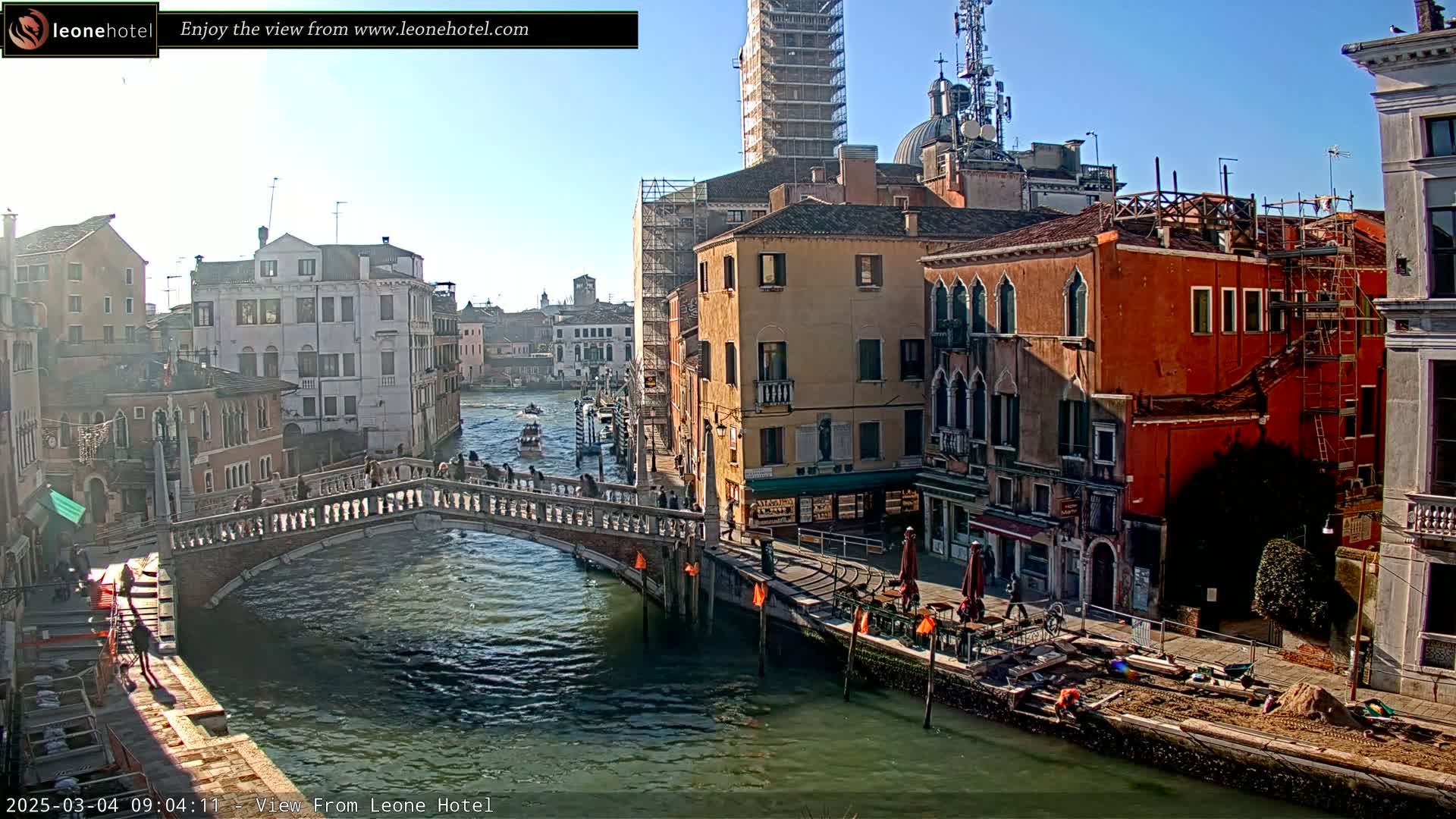 A sunny day in Venice shows a canal with a stone bridge, several multi-story buildings lining the waterway, and some boats on the water.