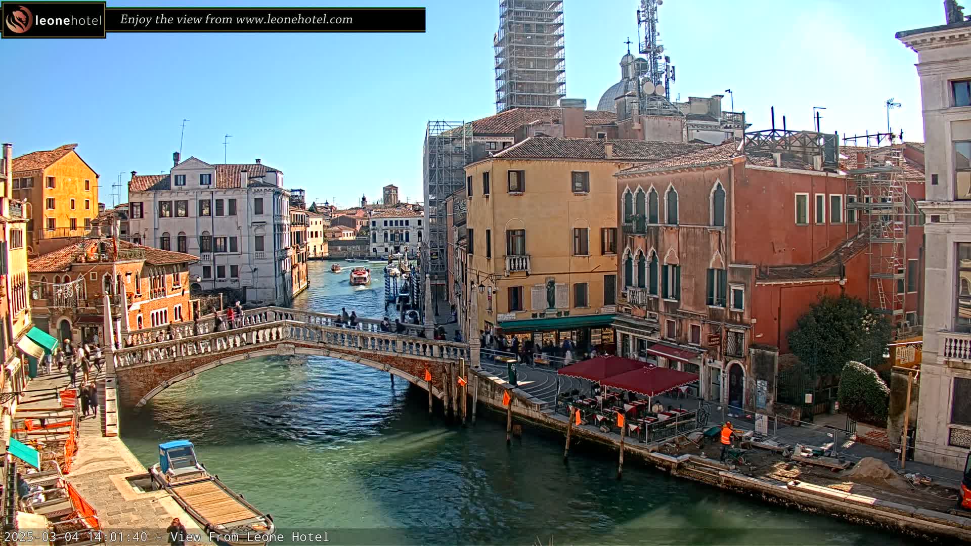 A sunny day in Venice shows a canal with boats, a stone bridge with pedestrians, and colorful buildings lining the waterway, some of which are under construction.