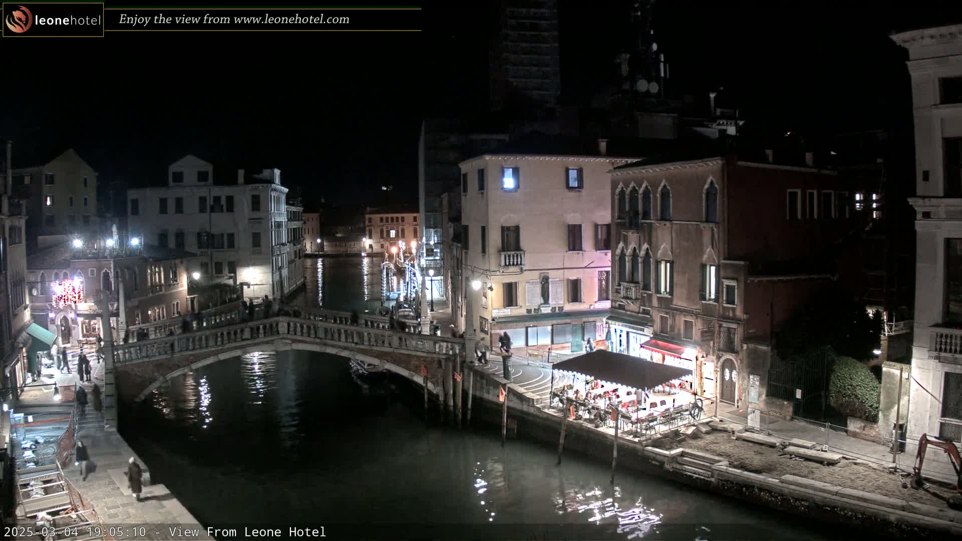 A nighttime view of a canal in Venice, Italy, shows several buildings lit along the waterway, a bridge with pedestrians crossing, and an outdoor restaurant with patrons seated under a canopy.