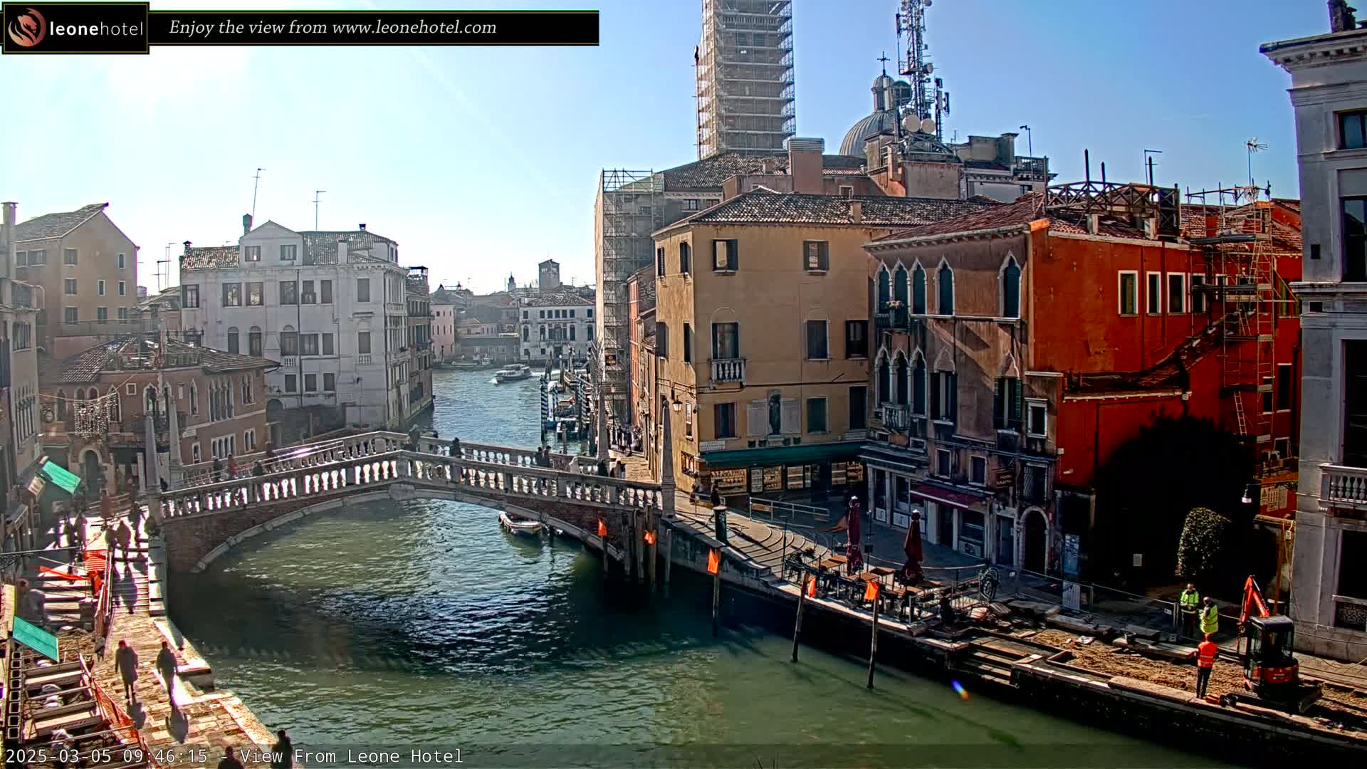 A sunny day in Venice shows a canal with a stone bridge, colorful buildings lining the waterway, and construction workers near a small excavator.
