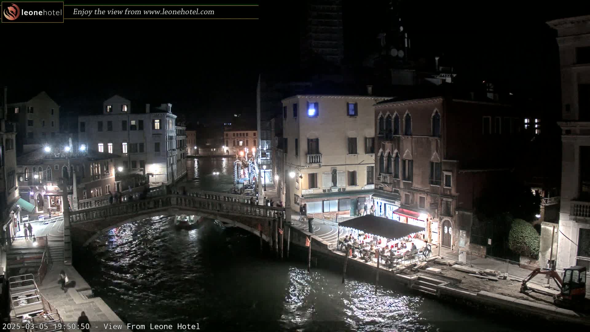 A nighttime view of a canal in Venice, showing bridges, buildings with lights, and a small outdoor restaurant along the water's edge.