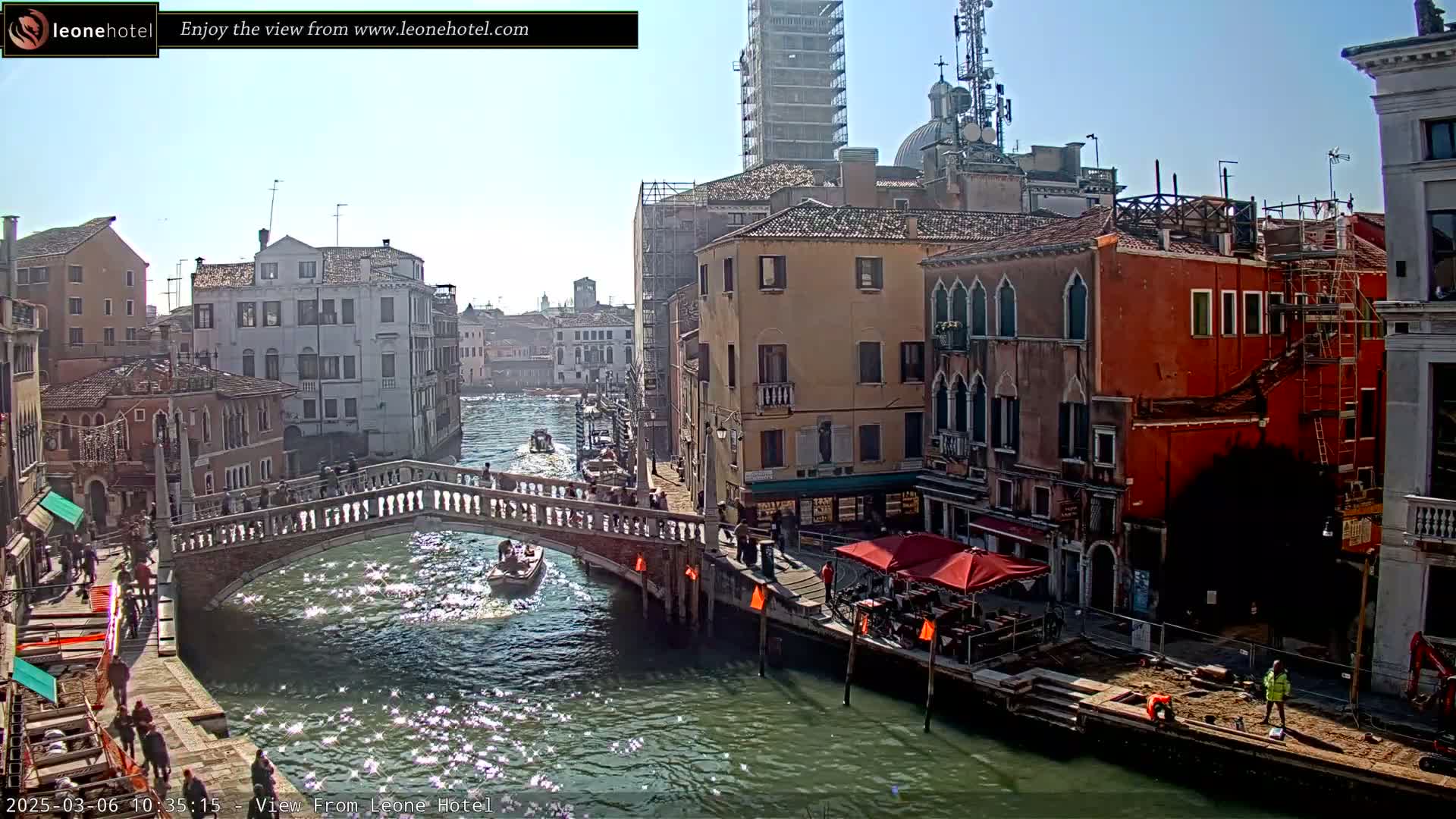 A sunny day in Venice shows a canal with gondolas and a stone bridge, flanked by colorful buildings, some undergoing construction.