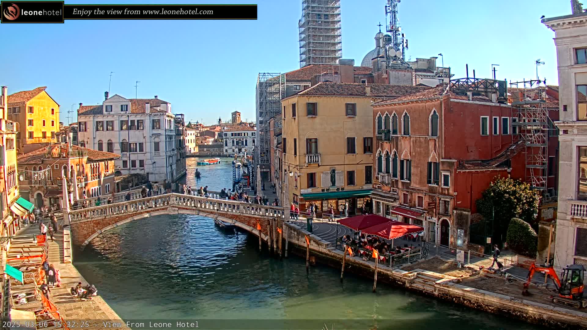 A sunny day in Venice shows a canal with a stone bridge, several buildings, and people walking along the canal bank.