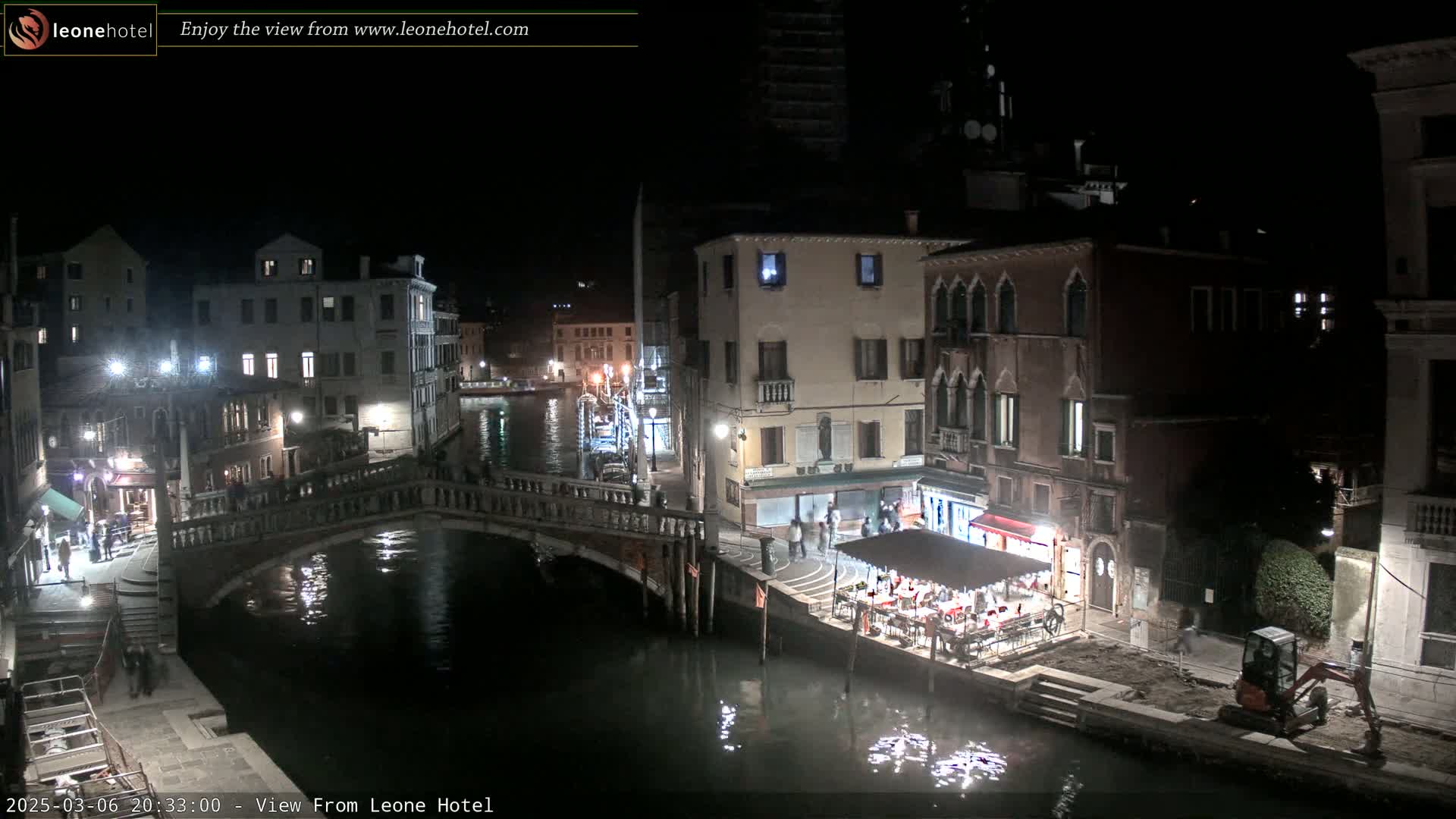 A nighttime view of a Venetian canal, showing bridges, buildings, and a small outdoor restaurant, with calm water reflecting the lights.
