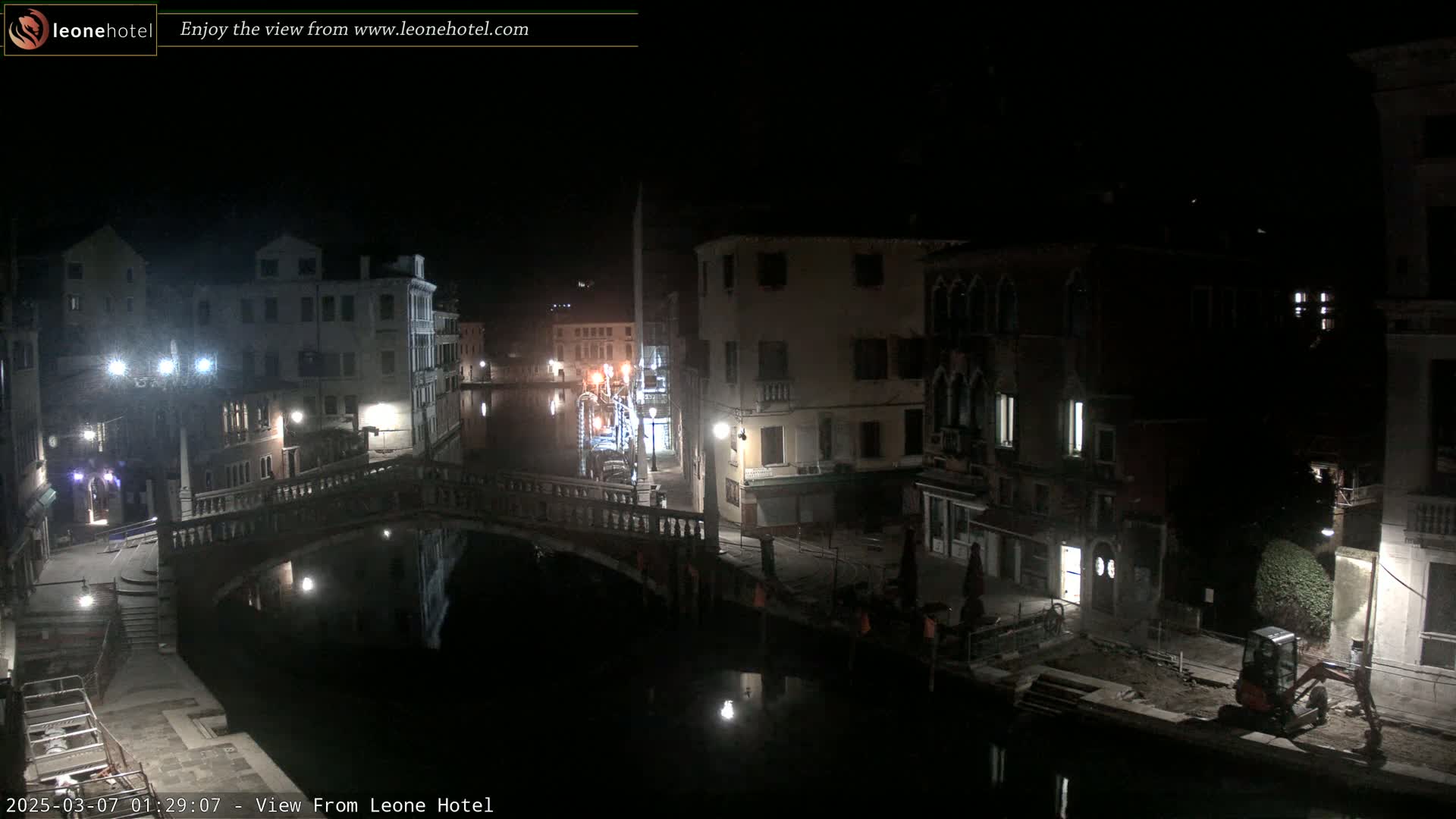 A nighttime view of a canal in Venice, Italy, shows several buildings lining the waterway, a bridge spanning the canal, and a small excavator parked near the water's edge.