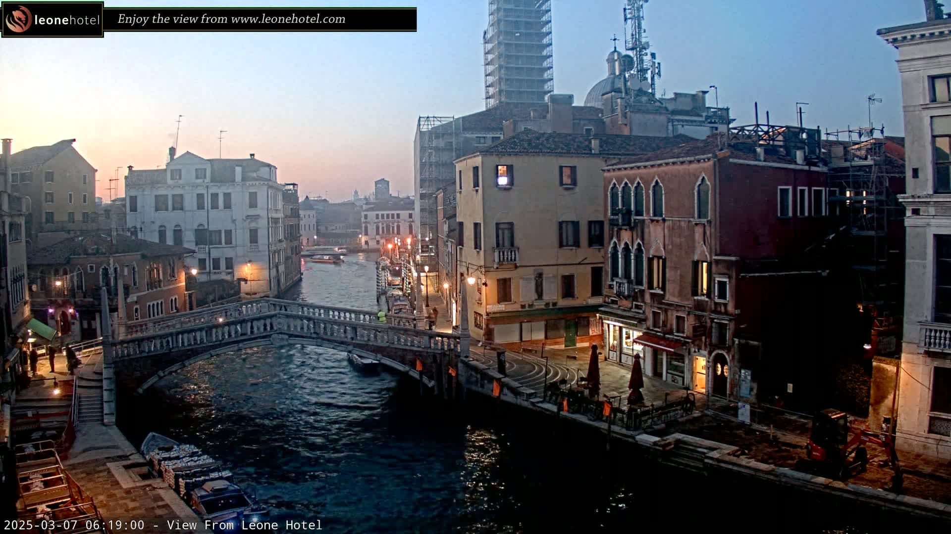 A twilight view of a canal in Venice, Italy, shows several buildings lining the waterway, a stone bridge, and a small boat on the water.