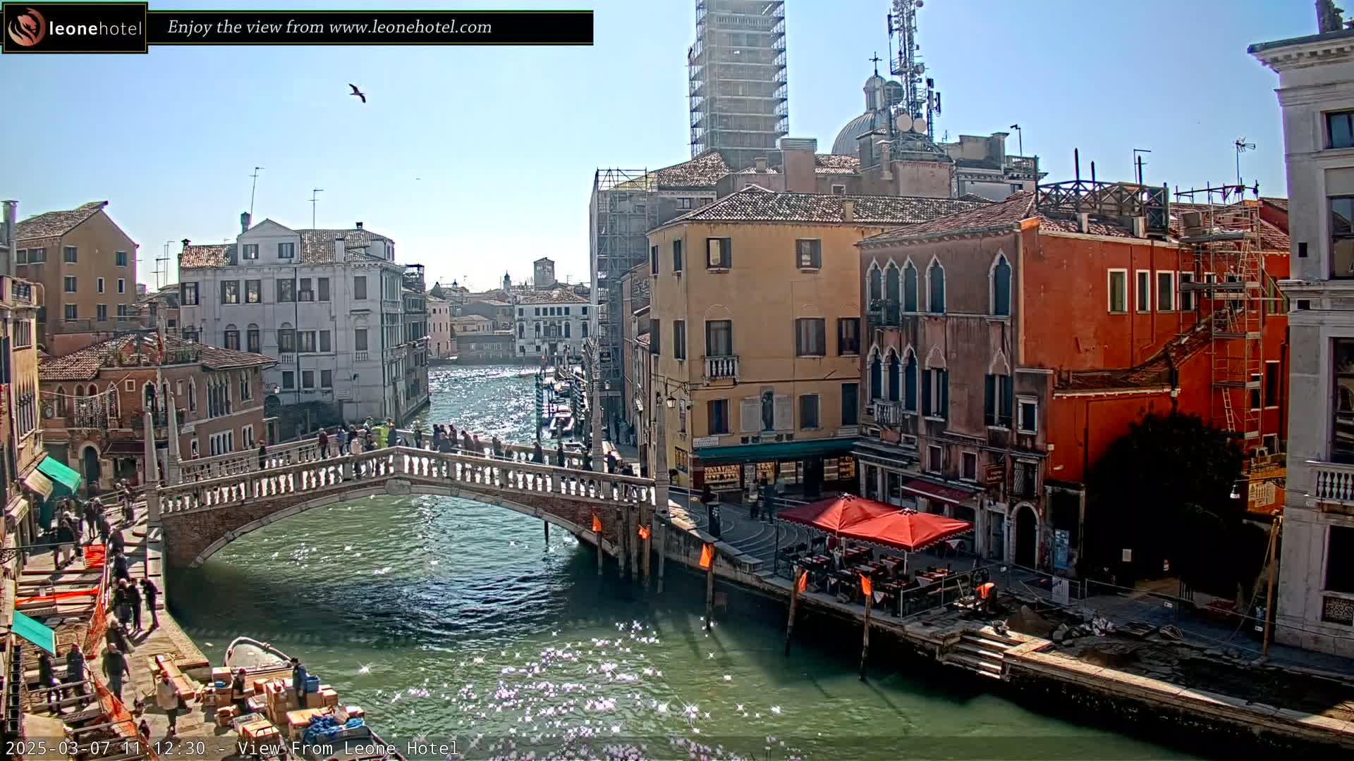 A sunny day in Venice shows a canal with a stone bridge crowded with people,  boats on the water, and colorful buildings lining the waterway.