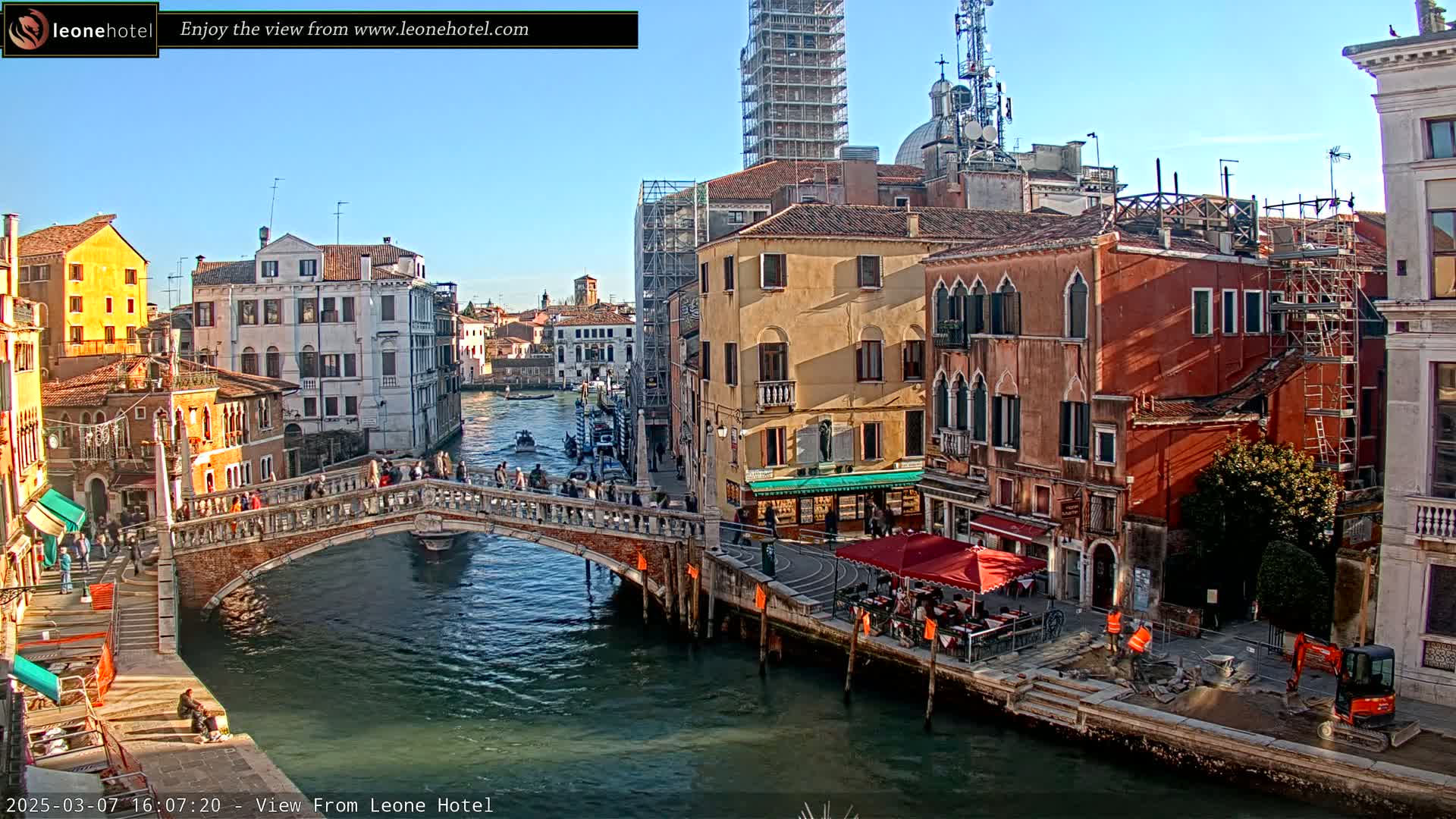 A sunny day in Venice shows a canal with a bridge, many pedestrians, several buildings, and construction taking place on the canal's edge.
