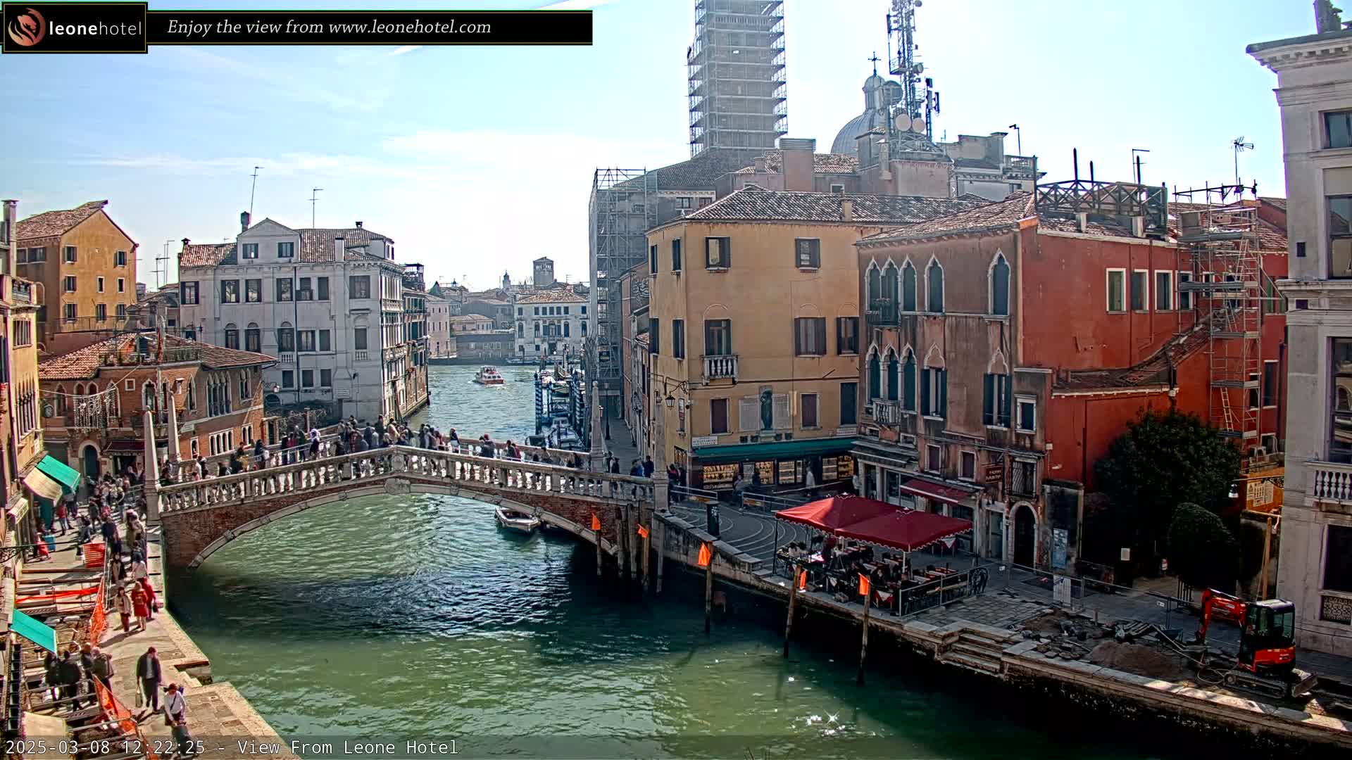A sunny day in Venice shows a canal with a stone bridge crowded with pedestrians, and colorful buildings lining the waterway, some under construction.