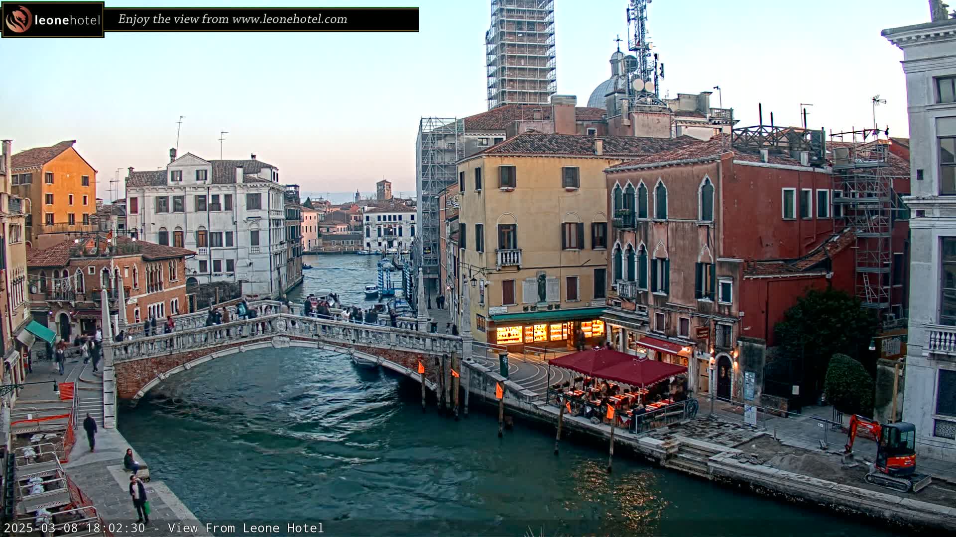 A stone bridge over a canal in Venice, Italy, at dusk, with pedestrians crossing it and buildings lining the waterways, some under construction.