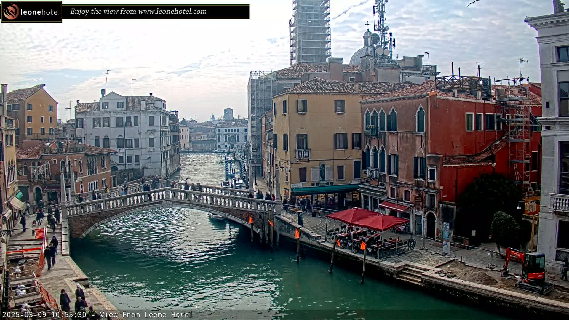 A stone bridge spans a canal in Venice, Italy, under a mostly cloudy sky, with colorful buildings lining the waterway and a small construction vehicle visible on the right.