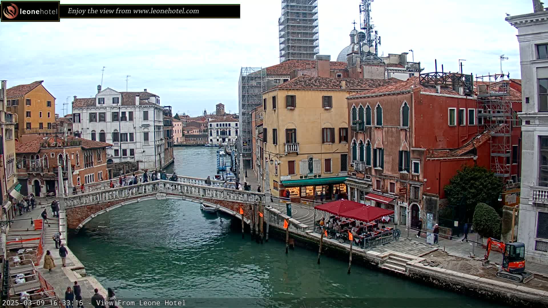 A stone bridge arches over a canal in Venice, Italy, under an overcast sky, with colorful buildings lining the waterway and scaffolding visible on some structures.