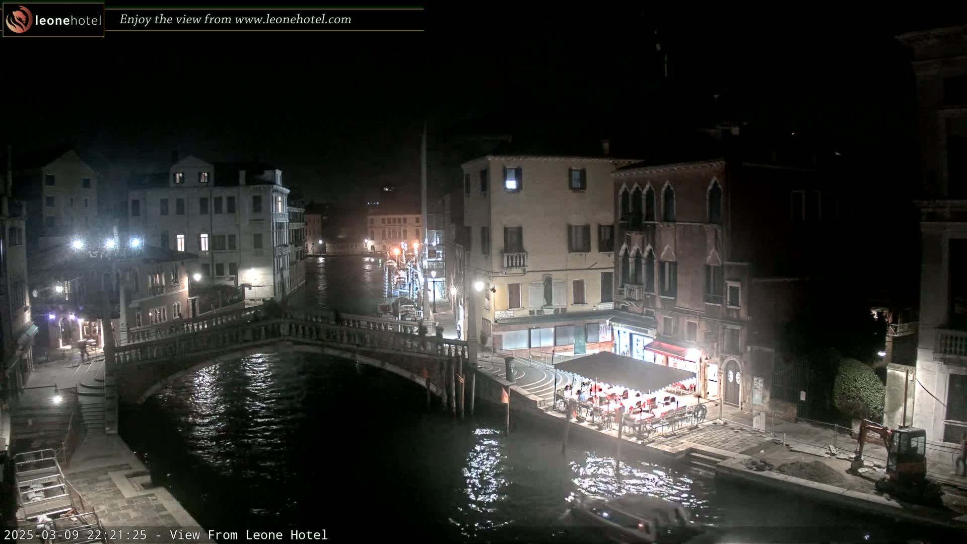 A nighttime view of a canal in Venice, with bridges, buildings, and an outdoor restaurant visible under a dark sky.
