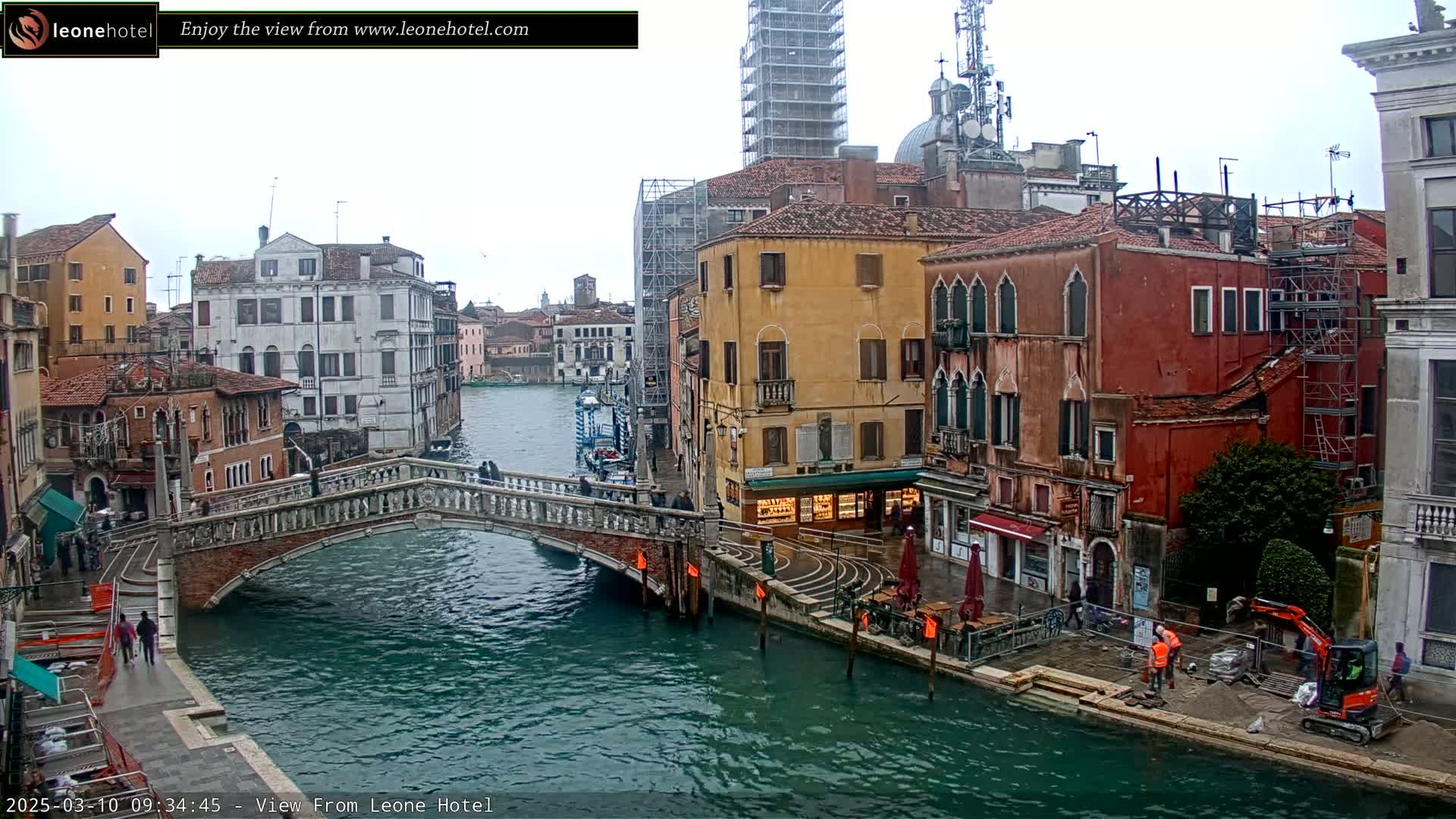 A view of a canal in Venice on an overcast day, showing a stone bridge, colorful buildings, and construction activity along the waterfront.