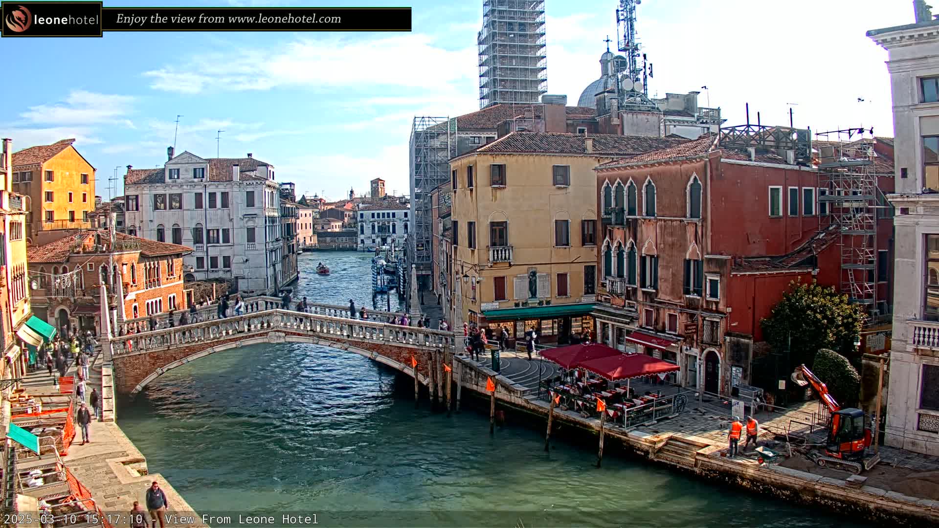 A sunny day in Venice shows a canal with a stone bridge, colorful buildings, pedestrians, gondolas, and construction workers near a small excavator.