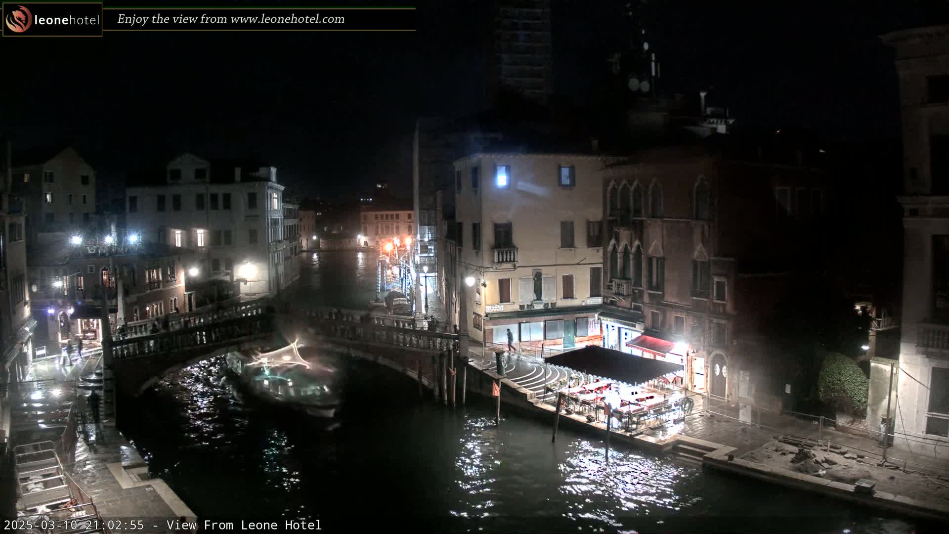A nighttime view of a canal in Venice, with several buildings illuminated along the waterway and a small boat docked near a restaurant.