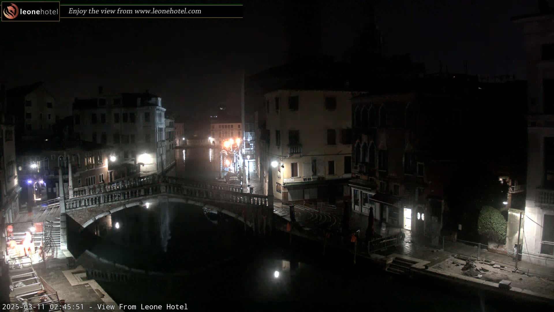 A nighttime view of a canal in Venice, Italy, shows a stone bridge over dark water with buildings lining both sides of the canal, and the area is dimly lit.