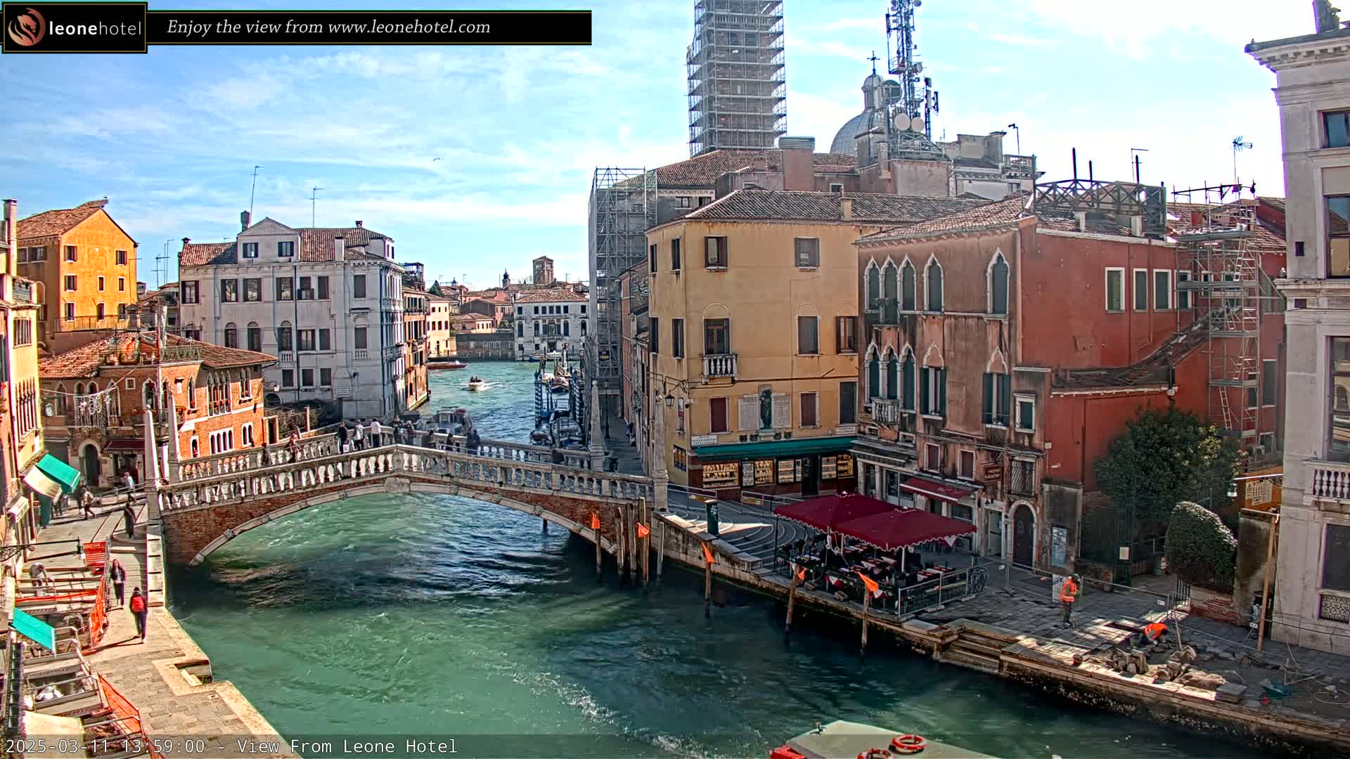 A sunny day in Venice shows a canal with a stone bridge, several buildings, and boats.