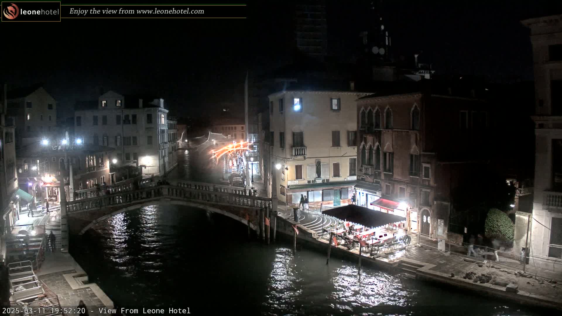 A nighttime view of a canal in Venice, showing several buildings, a bridge, and an outdoor restaurant, with calm water reflecting the lights.