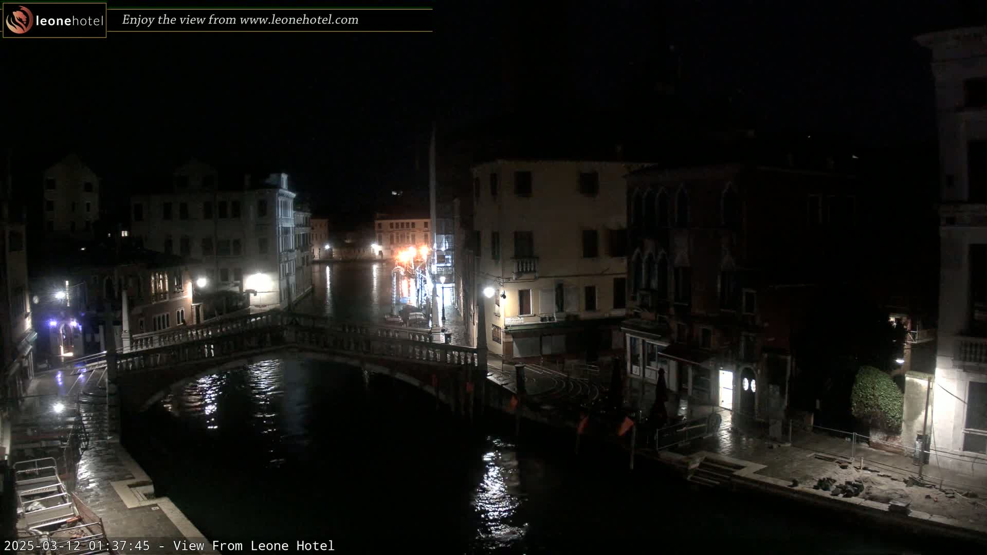 A nighttime view of a canal in Venice, showing a bridge and buildings lining the waterway under dark, possibly rainy conditions.