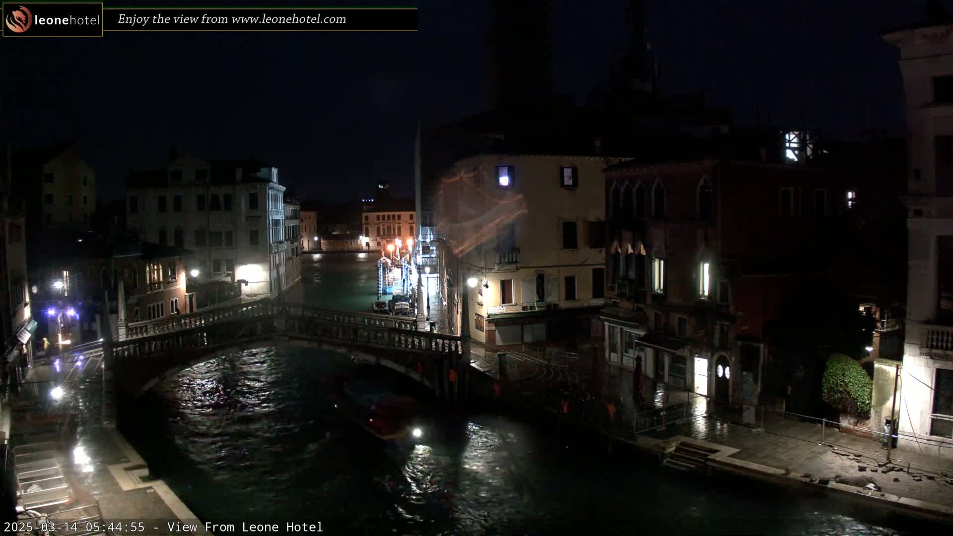 A nighttime view of a canal in Venice, Italy, showing bridges, buildings, and a boat, with wet pavement indicating rain.