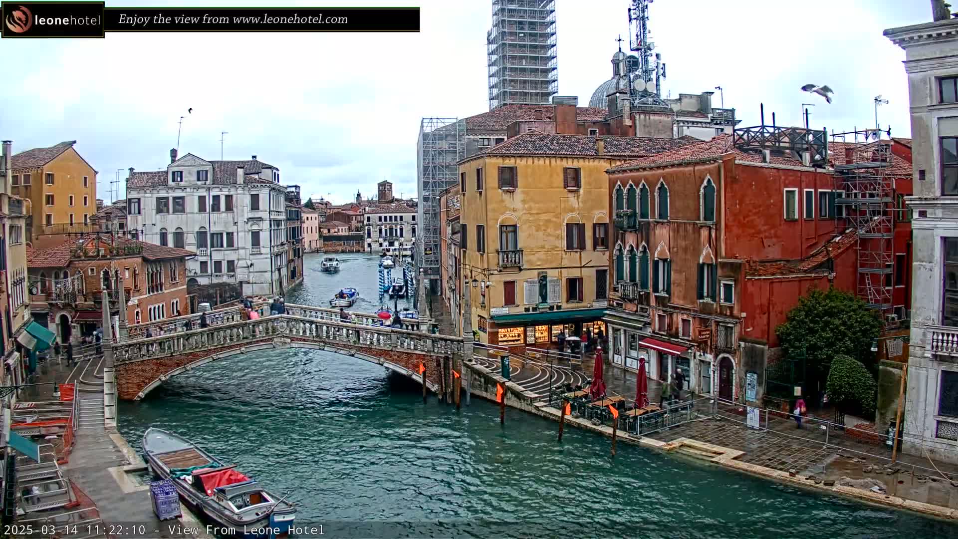 A view of a canal in Venice, Italy, on an overcast day, shows colorful buildings, a stone bridge, and several boats.