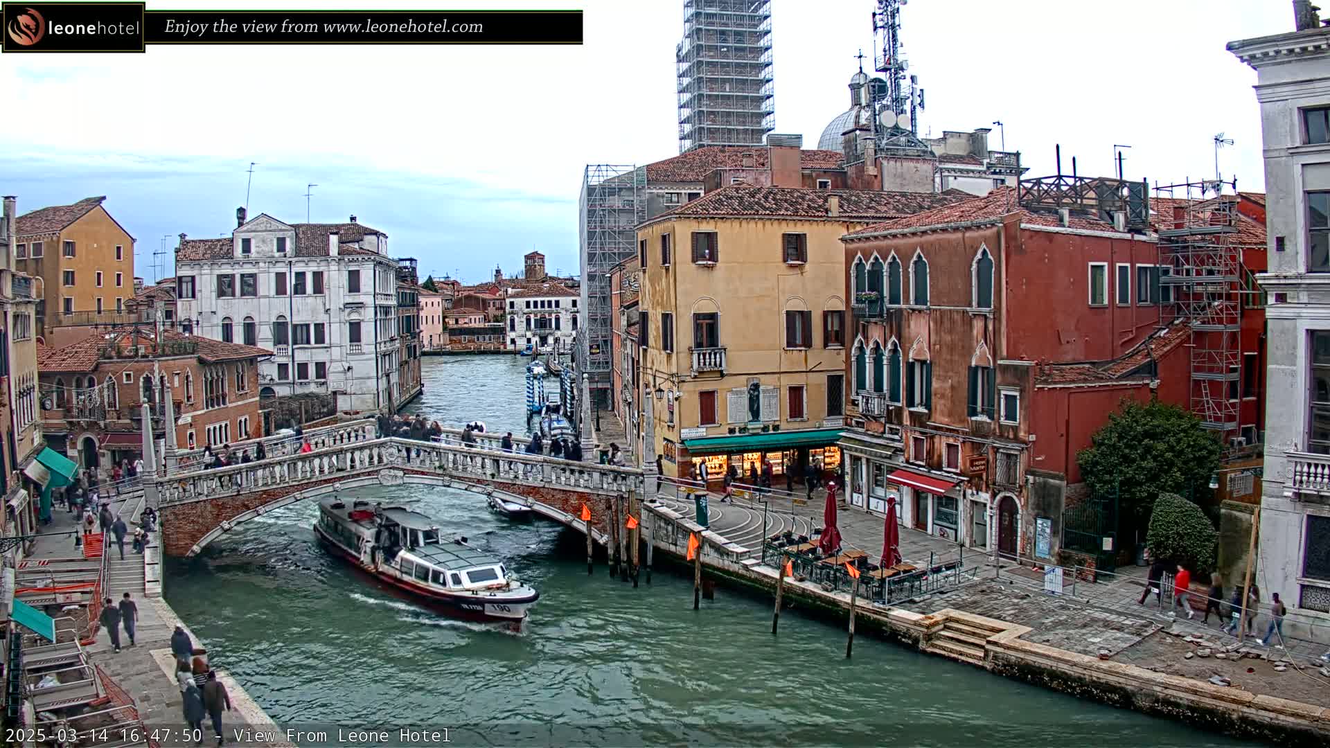 A waterway in Venice, Italy, is shown on an overcast day, with several boats passing under a bridge lined with pedestrians and buildings under construction.