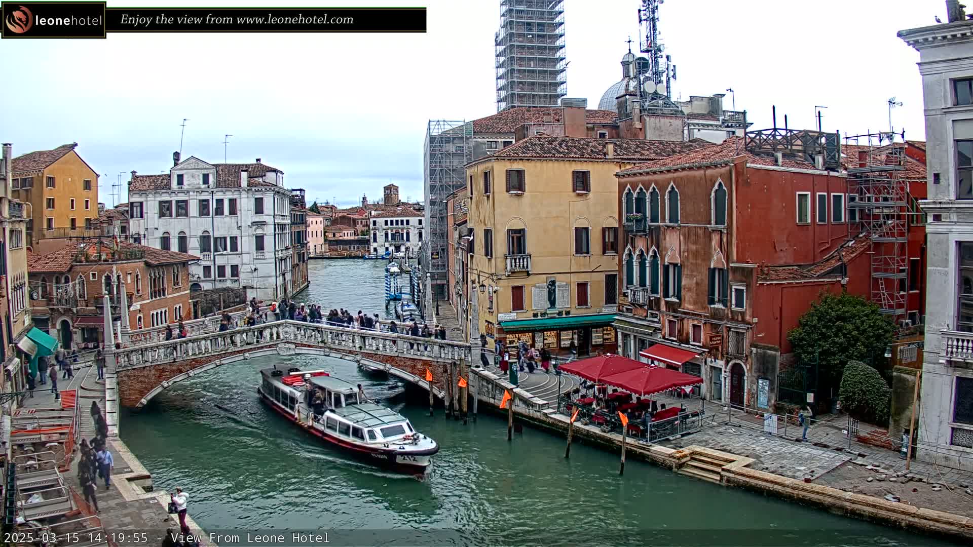 A canal in Venice, Italy, under an overcast sky, shows a bridge with people walking across it, a water taxi on the canal, and several buildings along the waterfront, some of which are under construction.