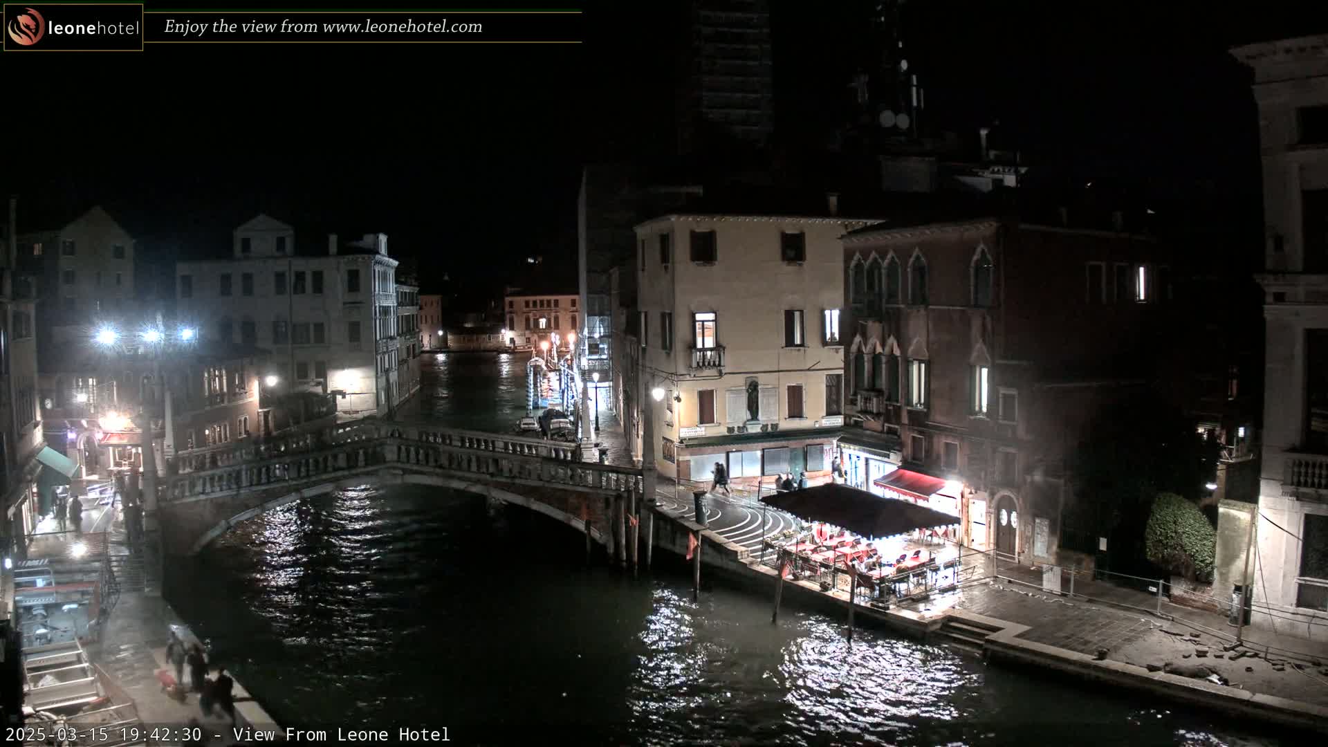 A nighttime view of a canal in Venice, Italy, showing a bridge, buildings, and a restaurant along the water's edge.
