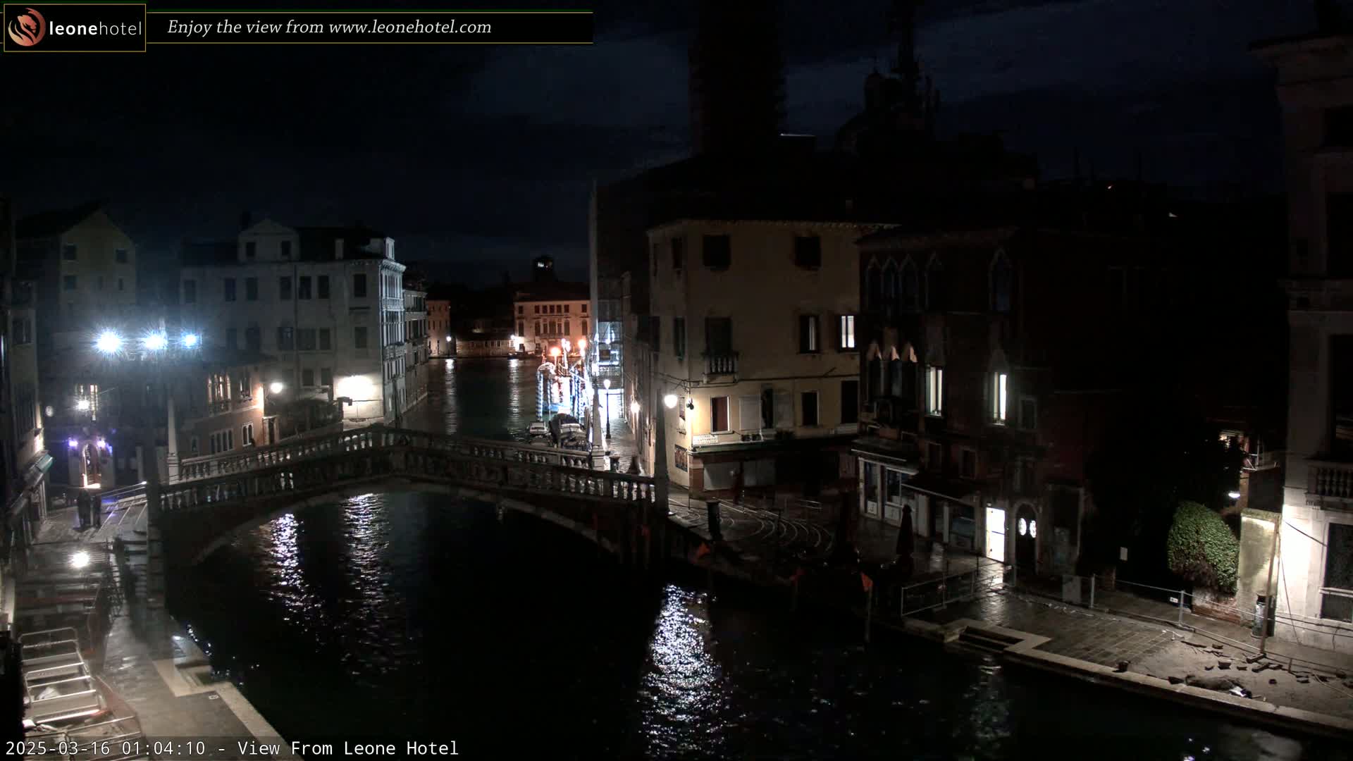 A nighttime view of a canal in Venice, Italy, shows a bridge, buildings lining the waterway, and a few people walking along the canal bank under dark, cloudy skies.