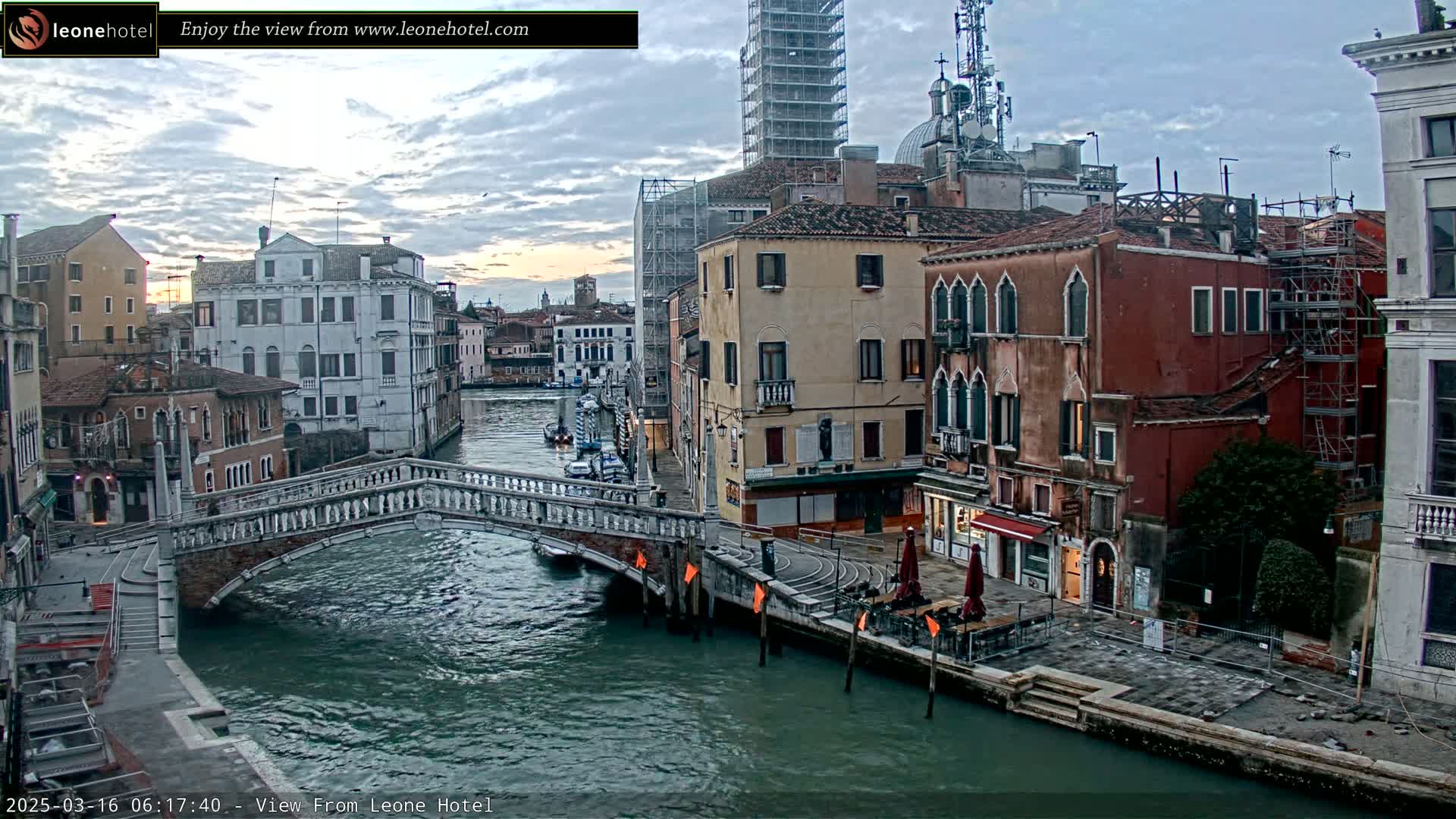 A stone bridge arches over a calm, dark-green canal in Venice, lined with historic buildings under a cloudy sky.