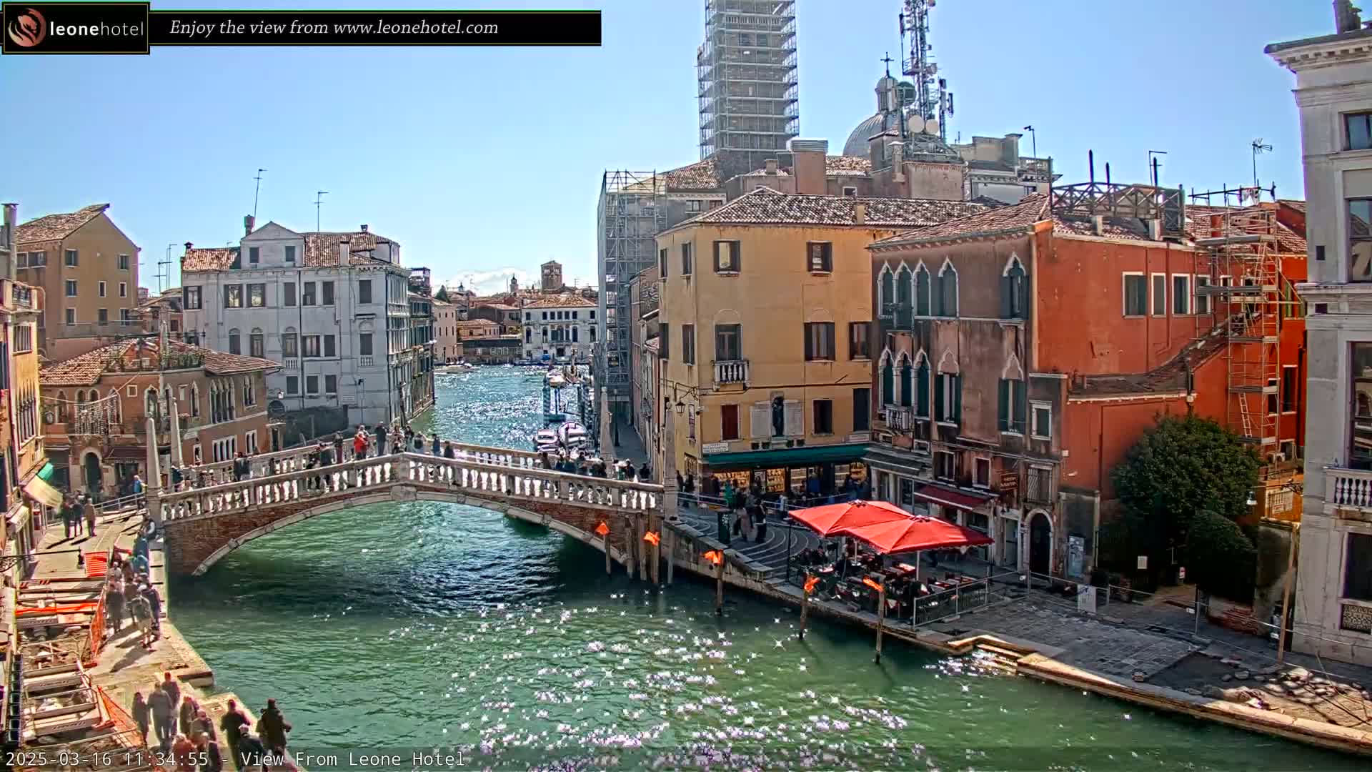 A sunny day in Venice shows a canal with a bridge, pedestrians, colorful buildings, and boats.