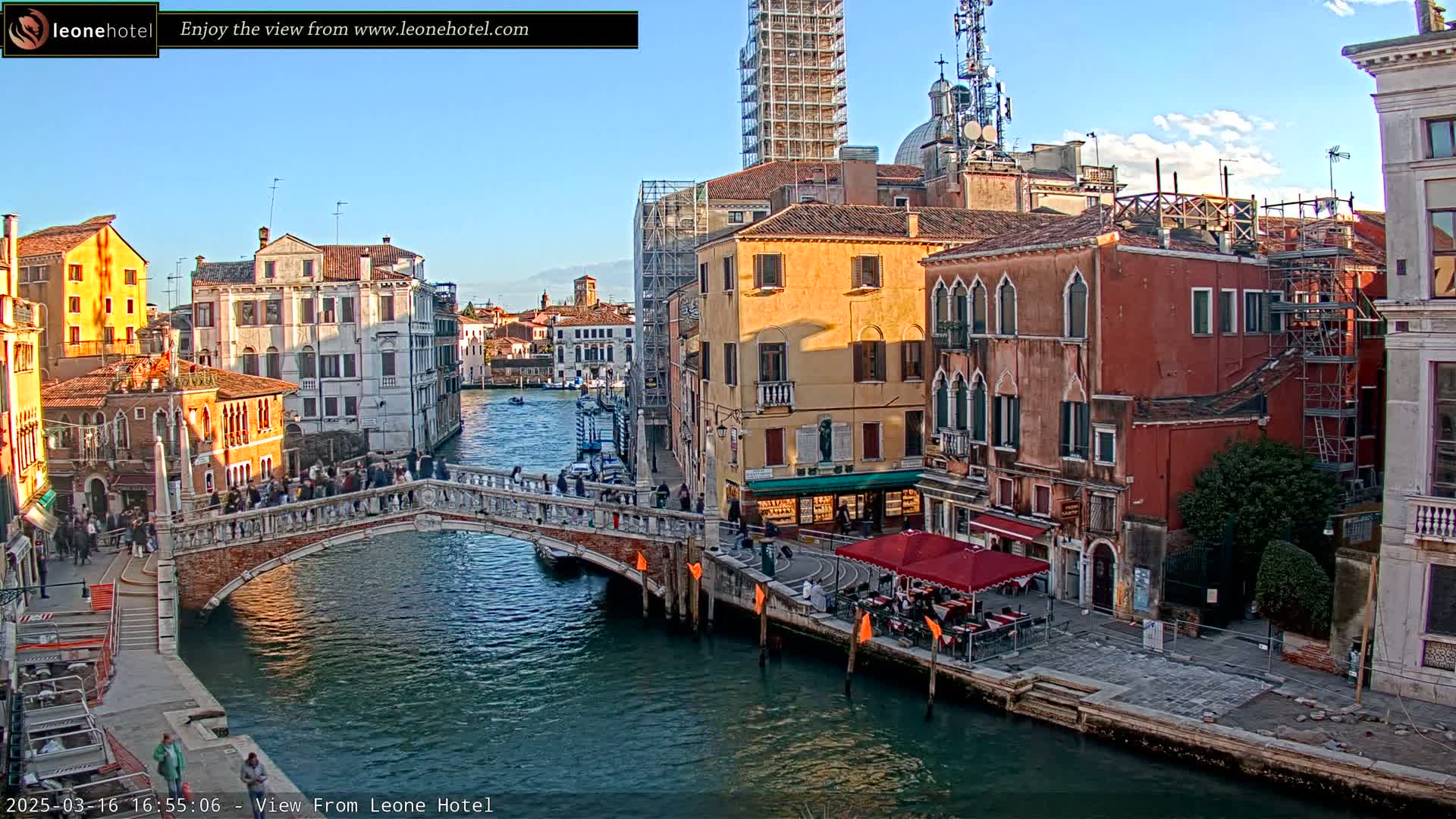 A Venetian canal scene with a bridge, colorful buildings, and people walking, under a mostly clear sky.