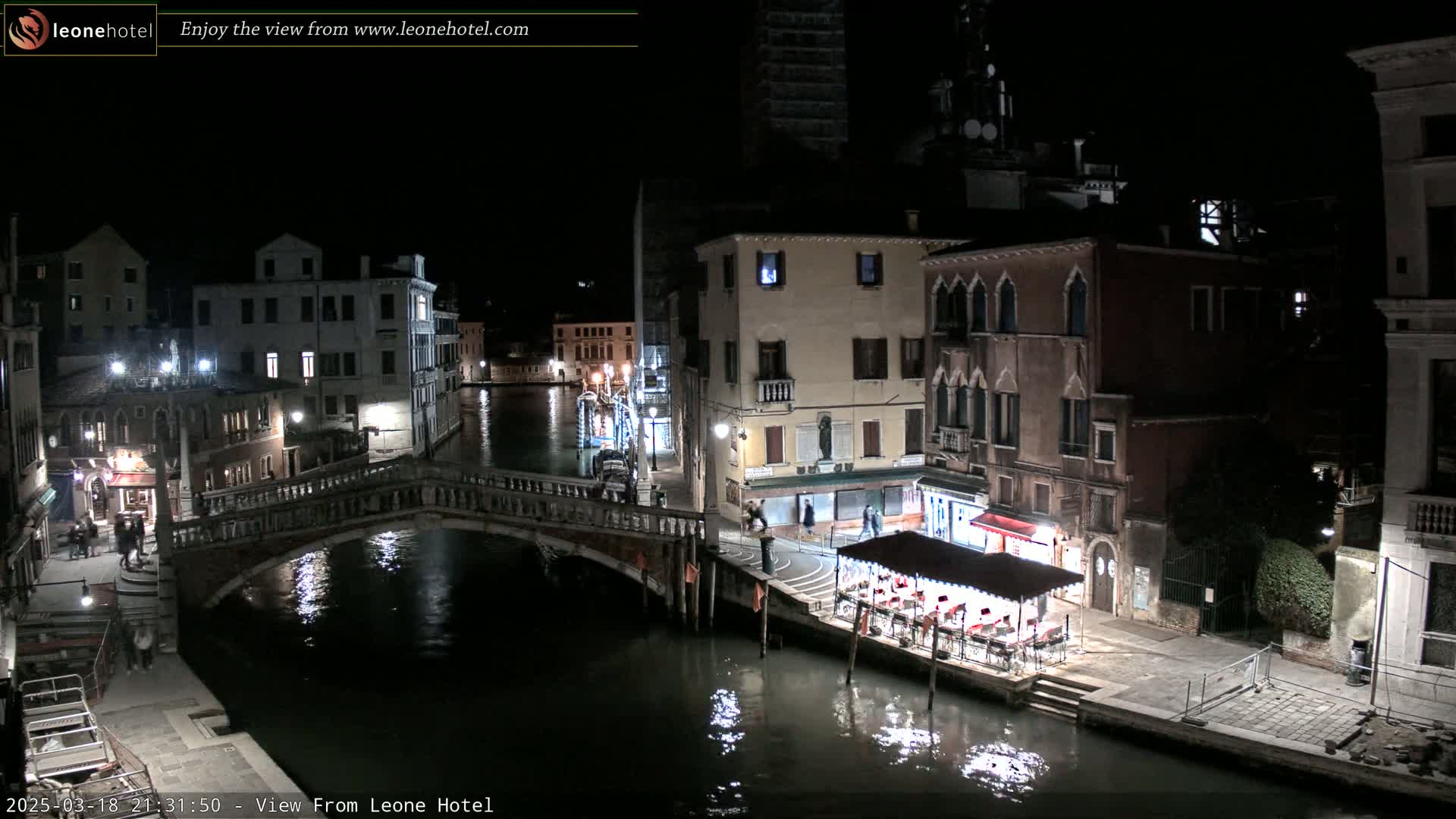 A nighttime view of a canal in Venice, Italy, shows a bridge, buildings lining the waterway, and an outdoor restaurant with patrons.