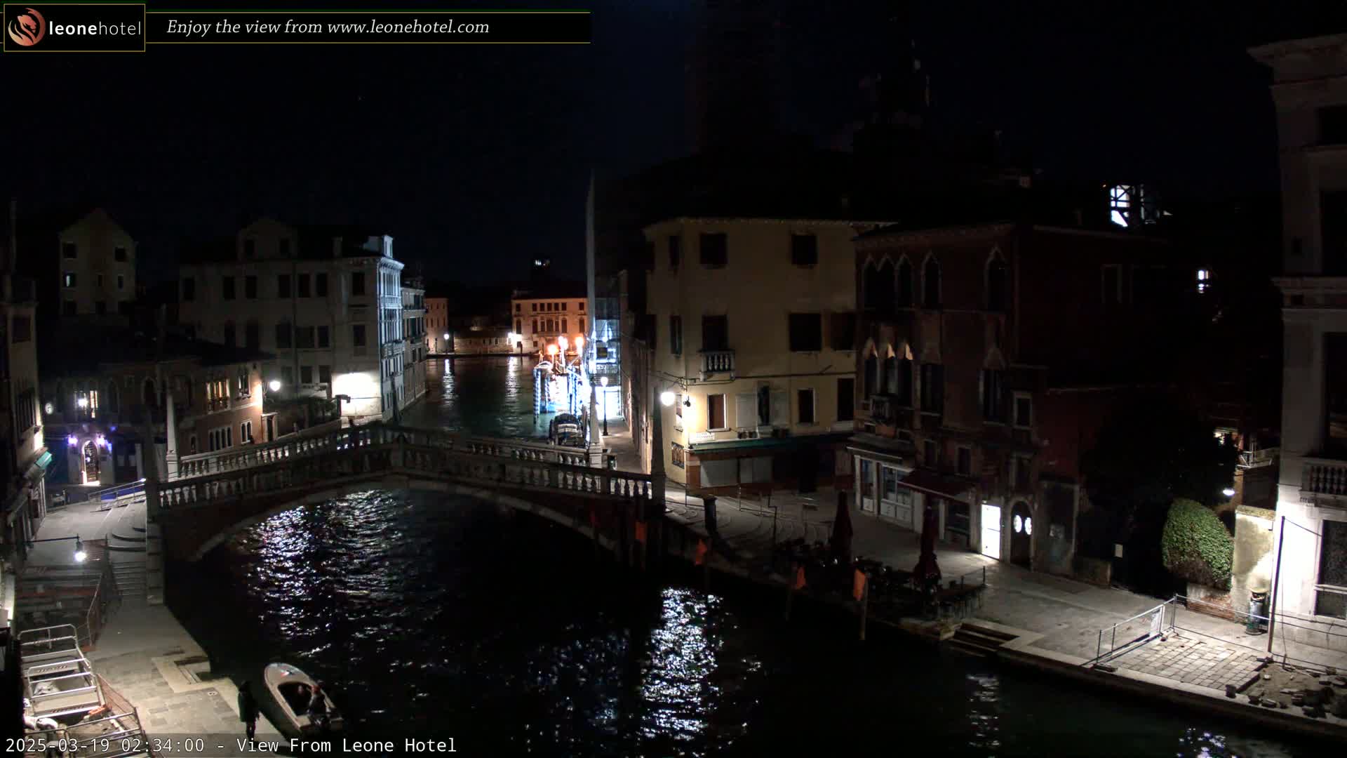 A nighttime view of a canal in Venice, Italy, shows a bridge, buildings with lights, and a small boat on the dark water.