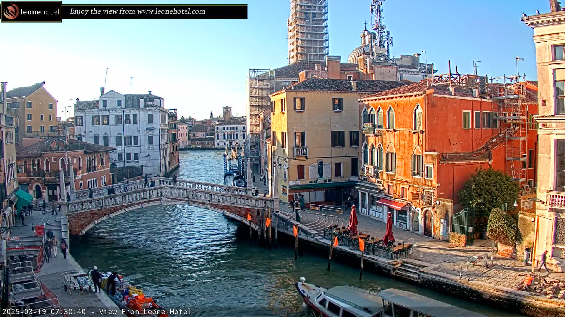 A canal in Venice, Italy, under a clear blue sky, features a stone bridge, colorful buildings, boats, and several pedestrians.