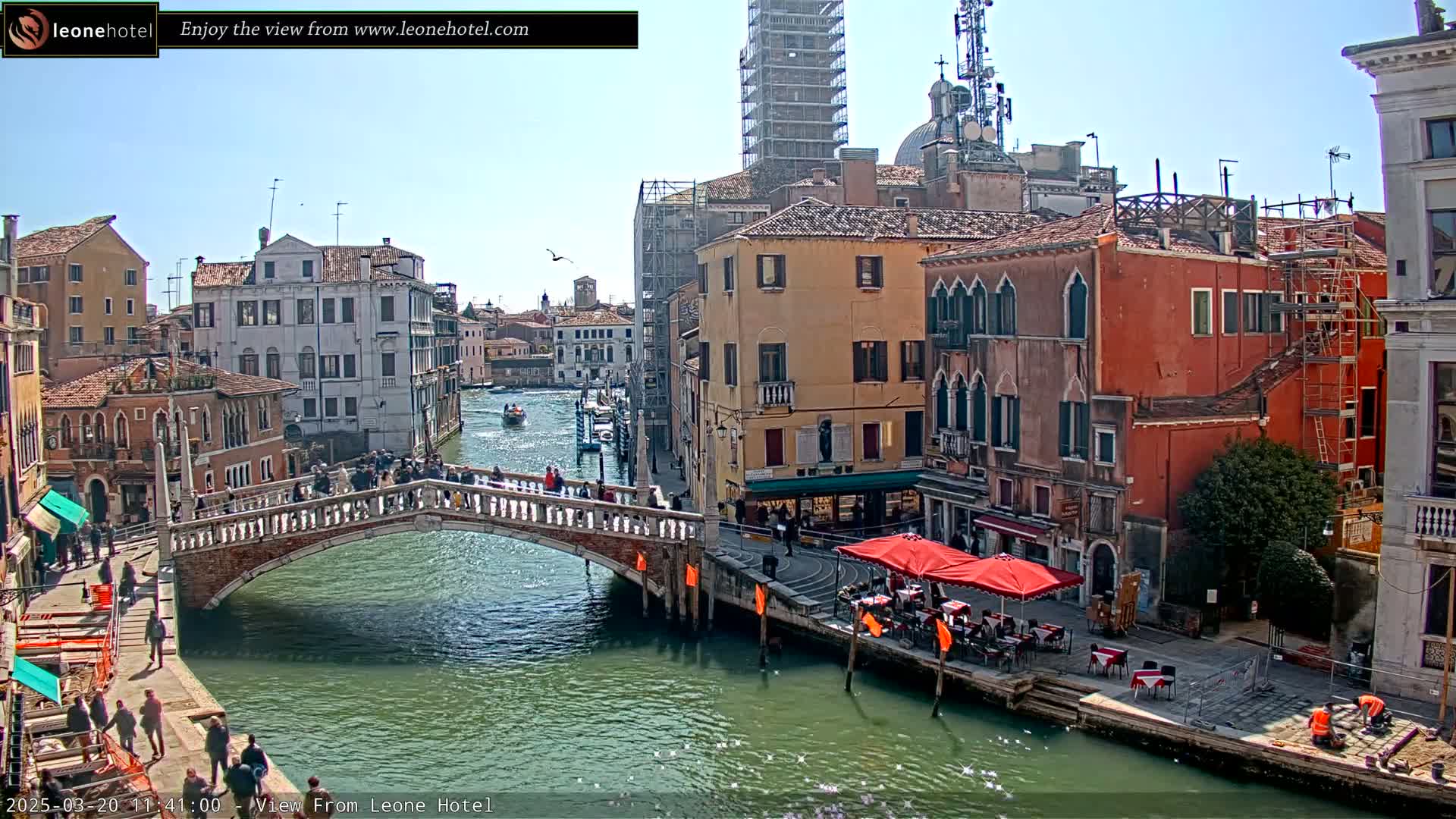 A sunny day in Venice shows a canal with a bridge, several buildings, and people walking on the bridge and along the canal-side.