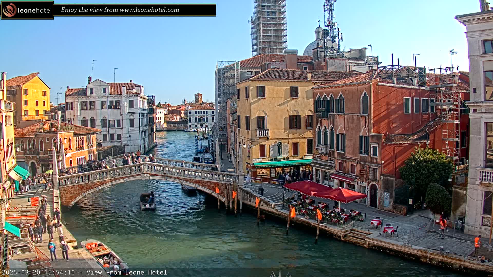 A sunny day in Venice shows a canal with a stone bridge, colorful buildings, and several people walking along the canal and bridge.
