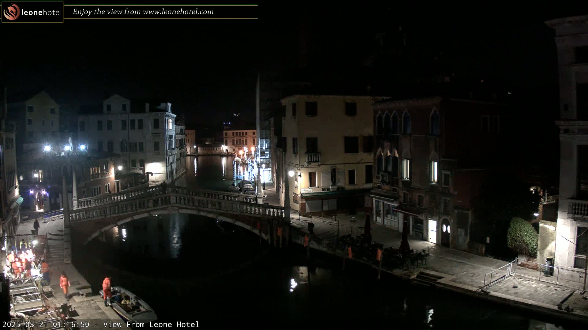 A nighttime view of a canal in Venice, Italy, with a bridge, buildings, and some people near the water's edge under dark, clear skies.