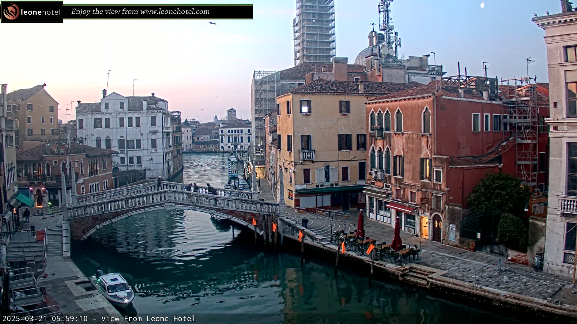 A tranquil canal scene in Venice, under a twilight sky, shows a stone bridge spanning the waterway lined with historic buildings, some under construction, with a few people strolling along the bridge and a small boat on the water.