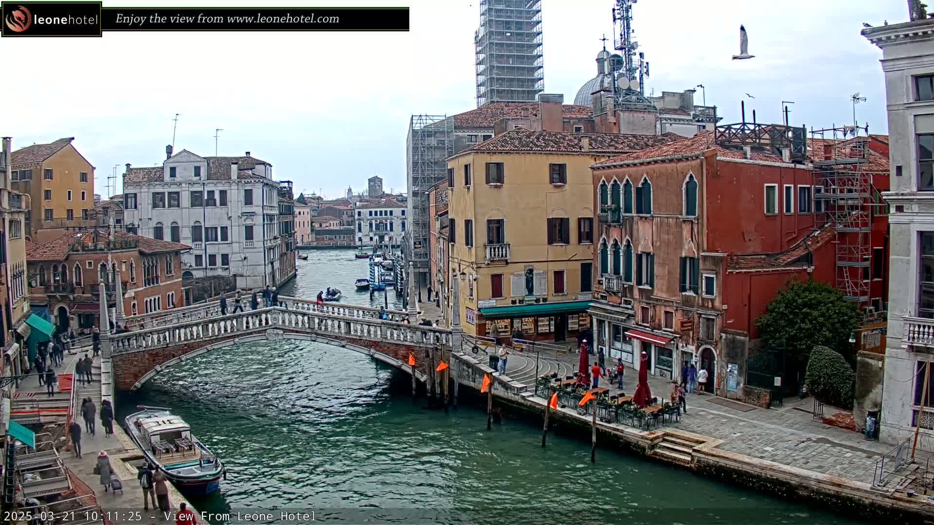 A canal in Venice, Italy, with several pedestrians on a bridge and along the waterfront, under an overcast sky.