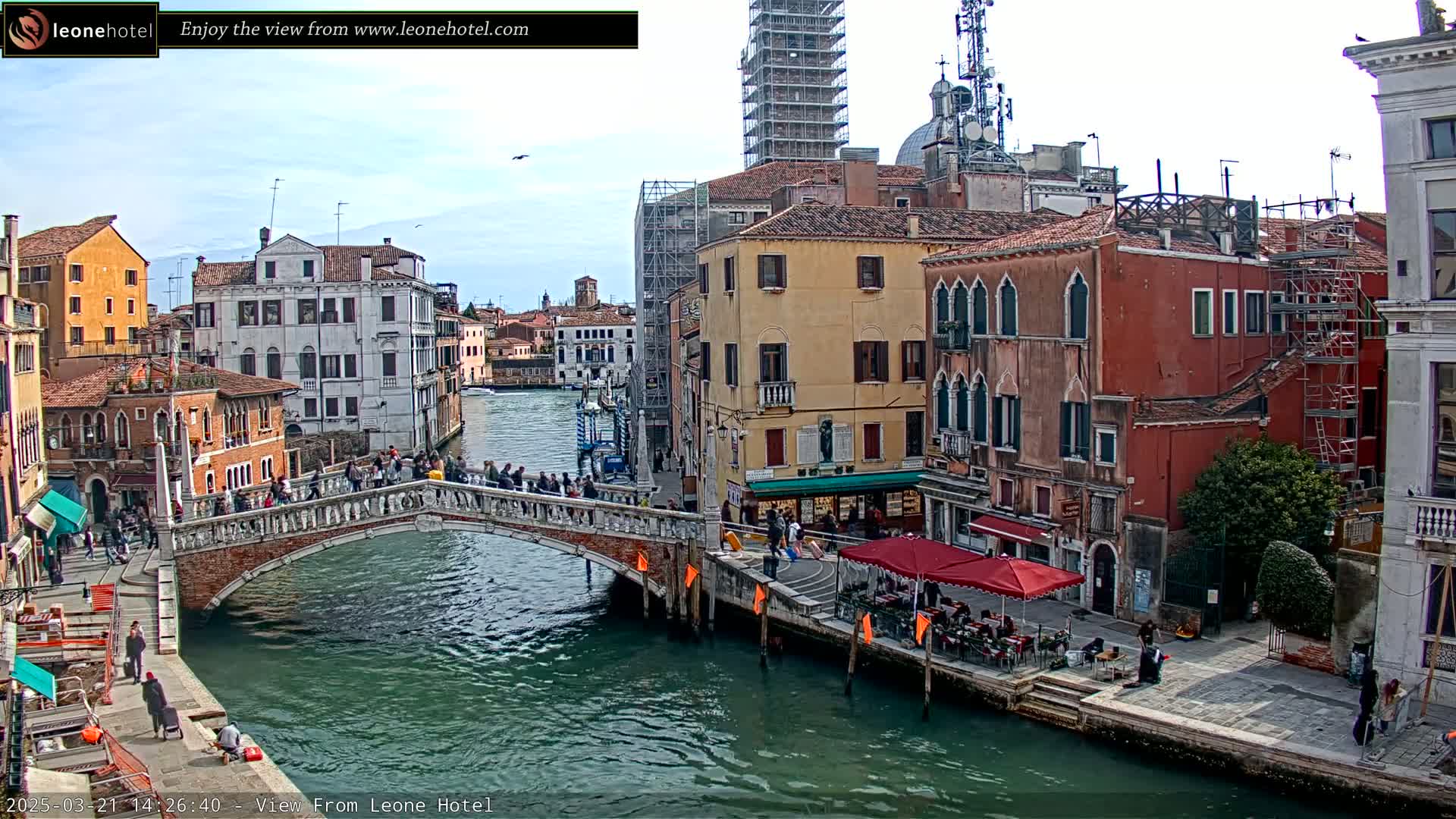 A bridge over a canal in Venice, Italy, under a mostly clear sky, is surrounded by several multi-story buildings, some of which are under construction.