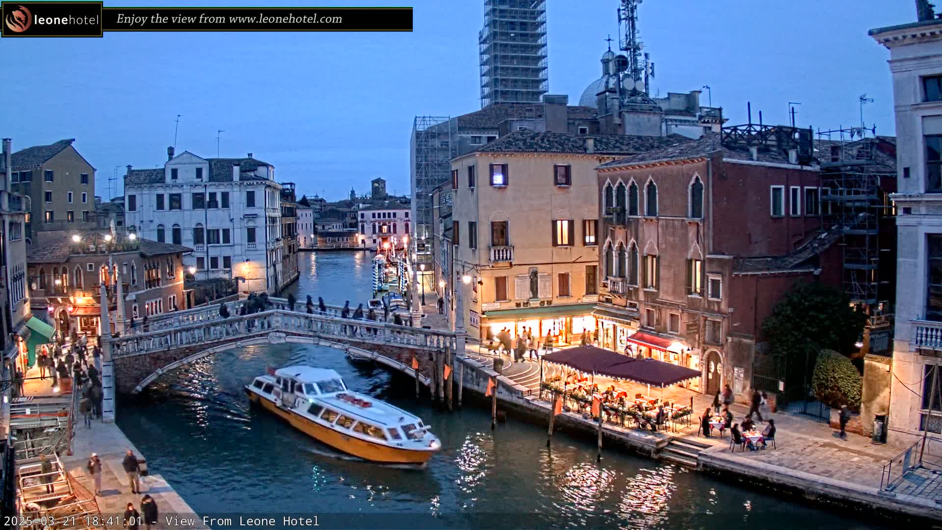 A canal in Venice at twilight, with a bridge, boats, buildings, and people along the waterways.