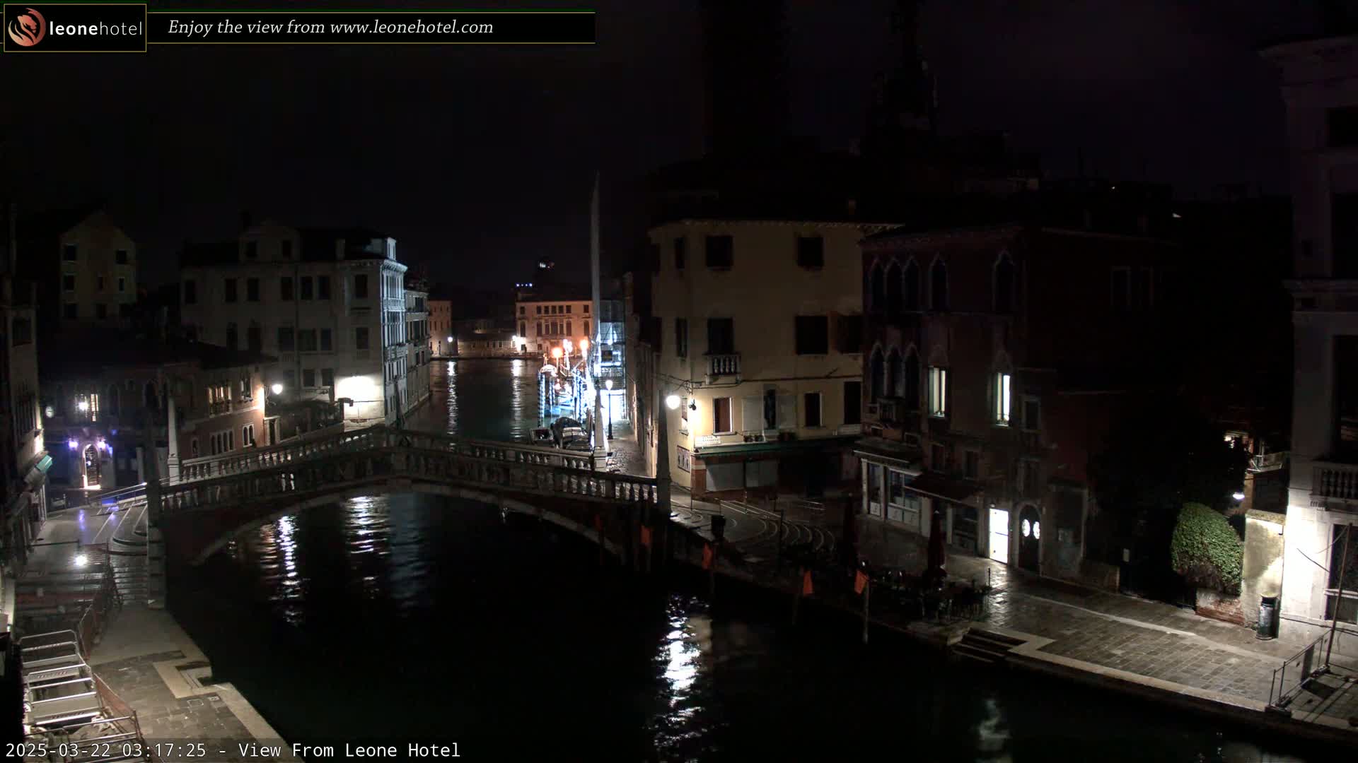 A nighttime view of a canal in Venice, Italy, shows a bridge and buildings illuminated by streetlights under a dark sky with wet pavement.