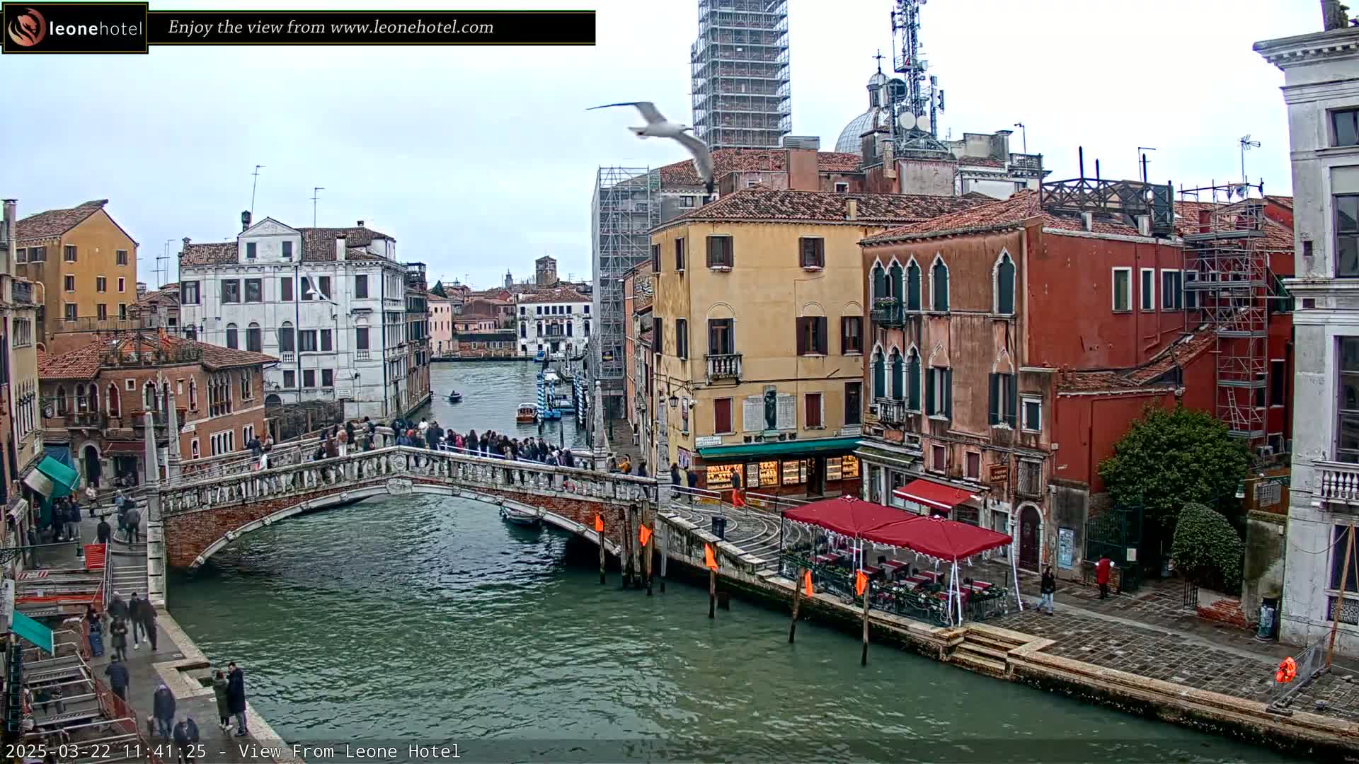 A stone bridge over a canal in Venice, Italy, with several buildings lining the waterway under an overcast sky, and a bird flying overhead.