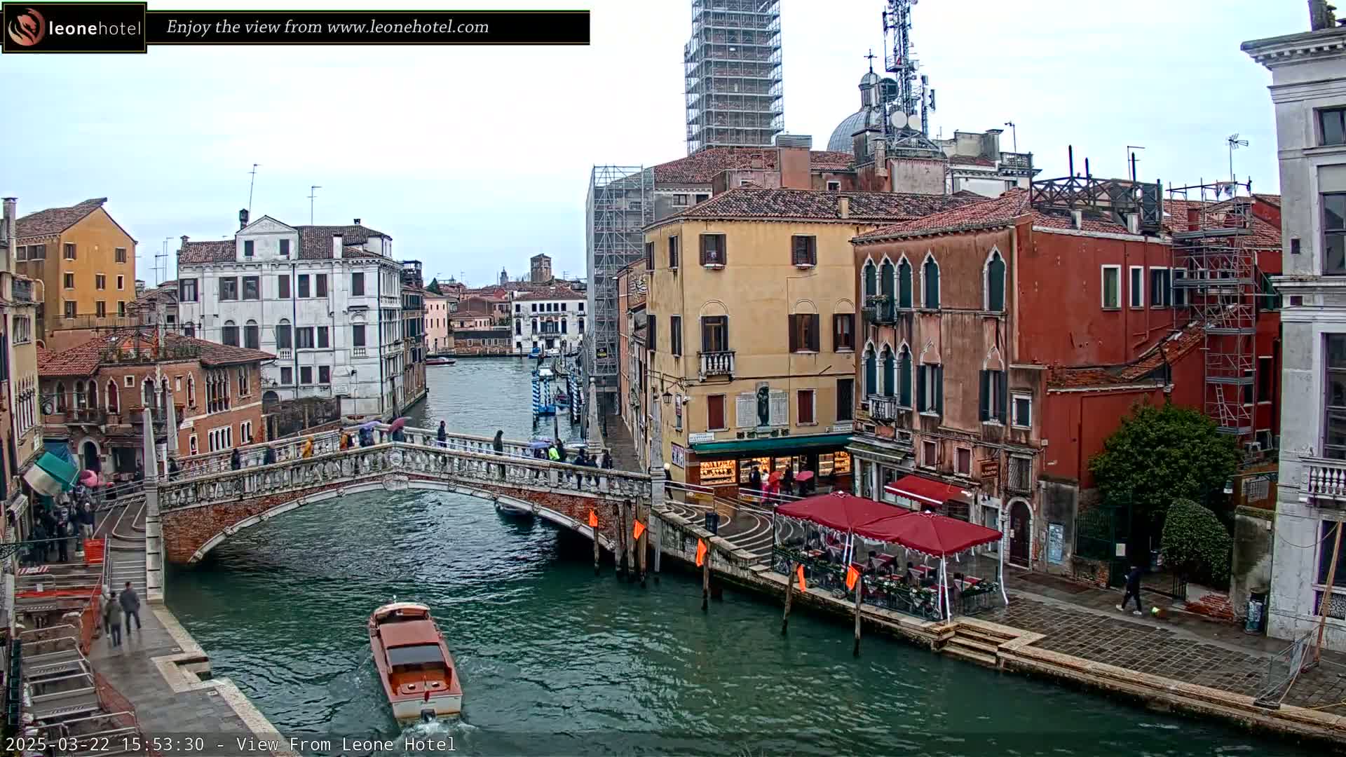 A canal in Venice, Italy, with pedestrians on a bridge and boats in the water, under an overcast sky.