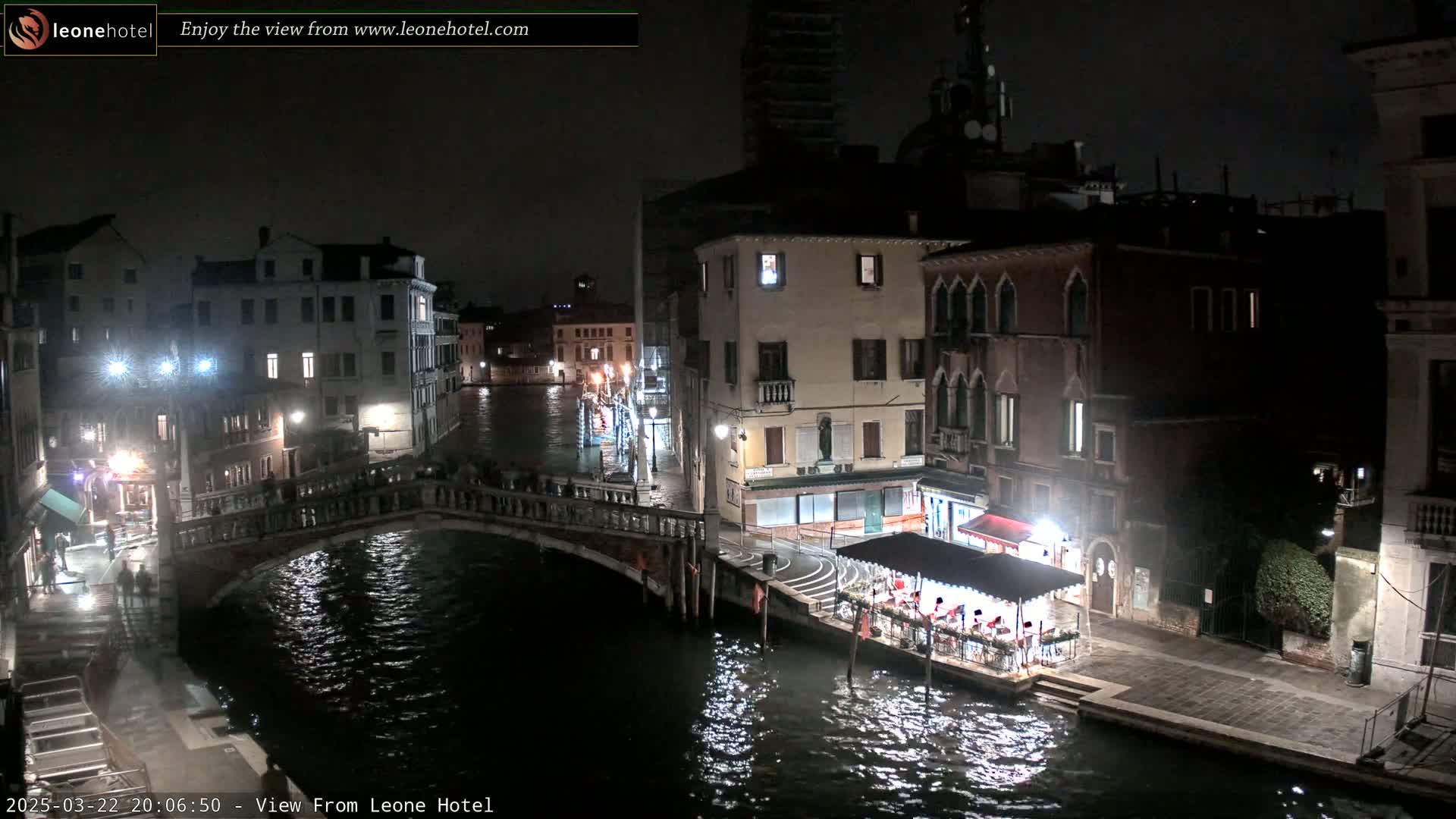 A nighttime view of a Venetian canal, showing bridges, buildings, and a restaurant on a boat, under a dark sky.